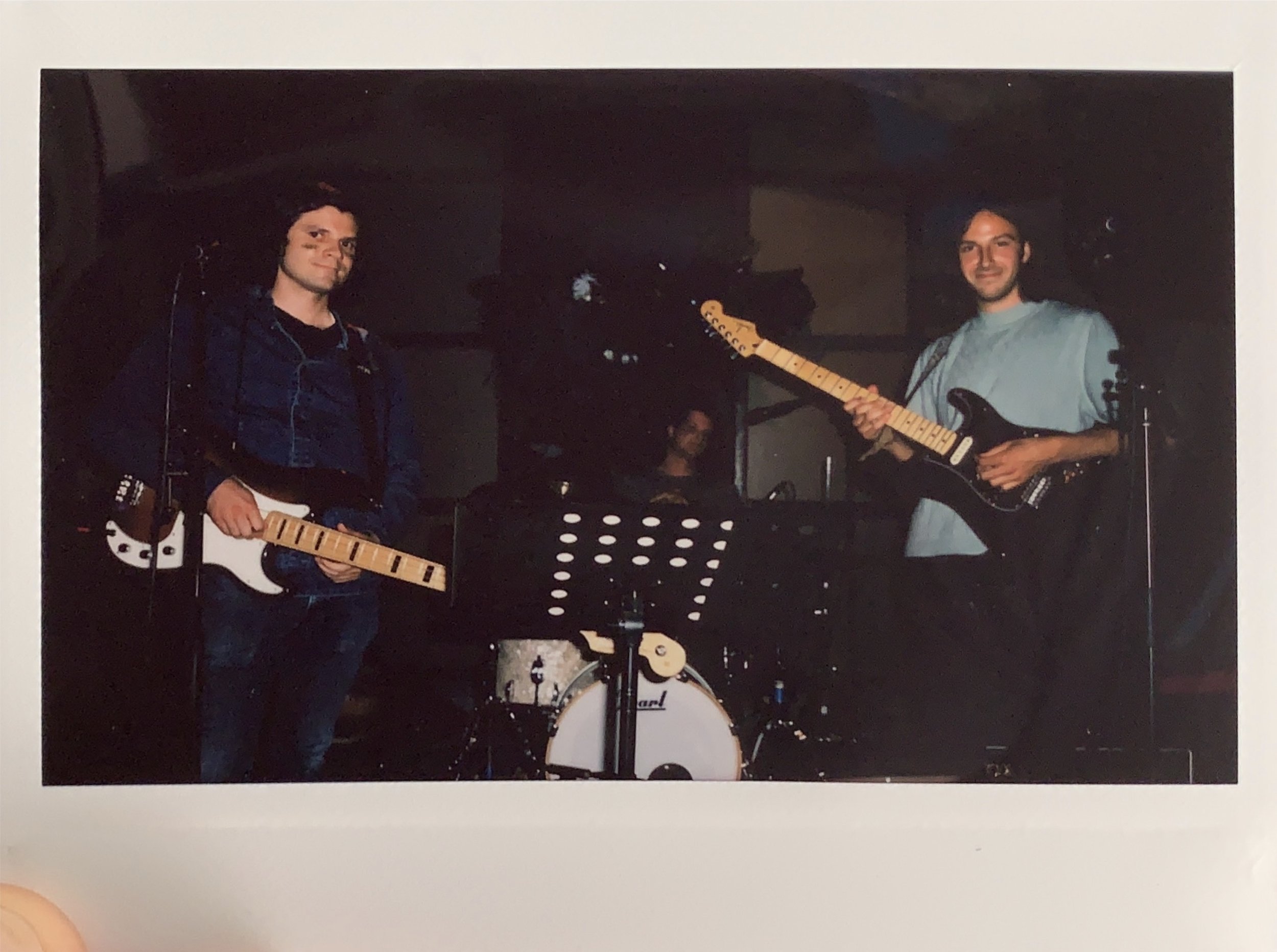 Three musicians with electric guitars and a drum set in a dark indoor setting.