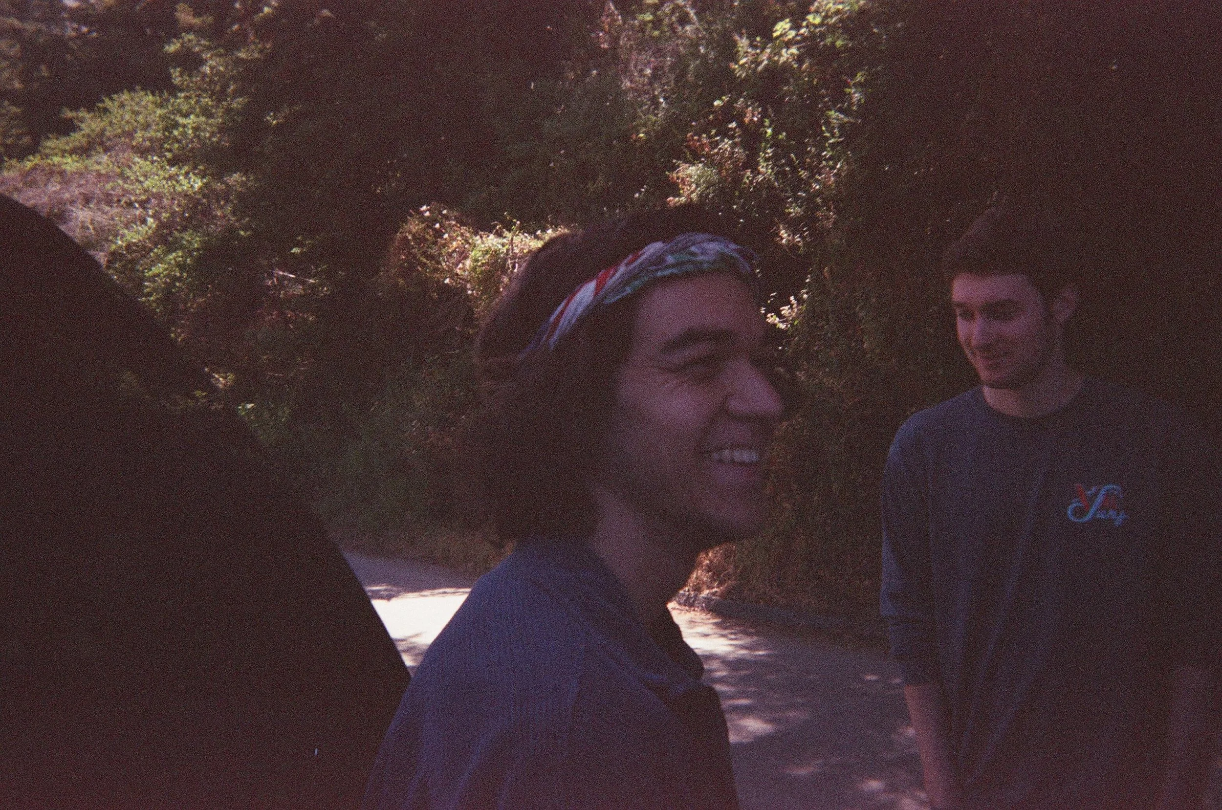 Two young men outdoors on a sunny day, standing on a tree-lined path, smiling and talking to each other.