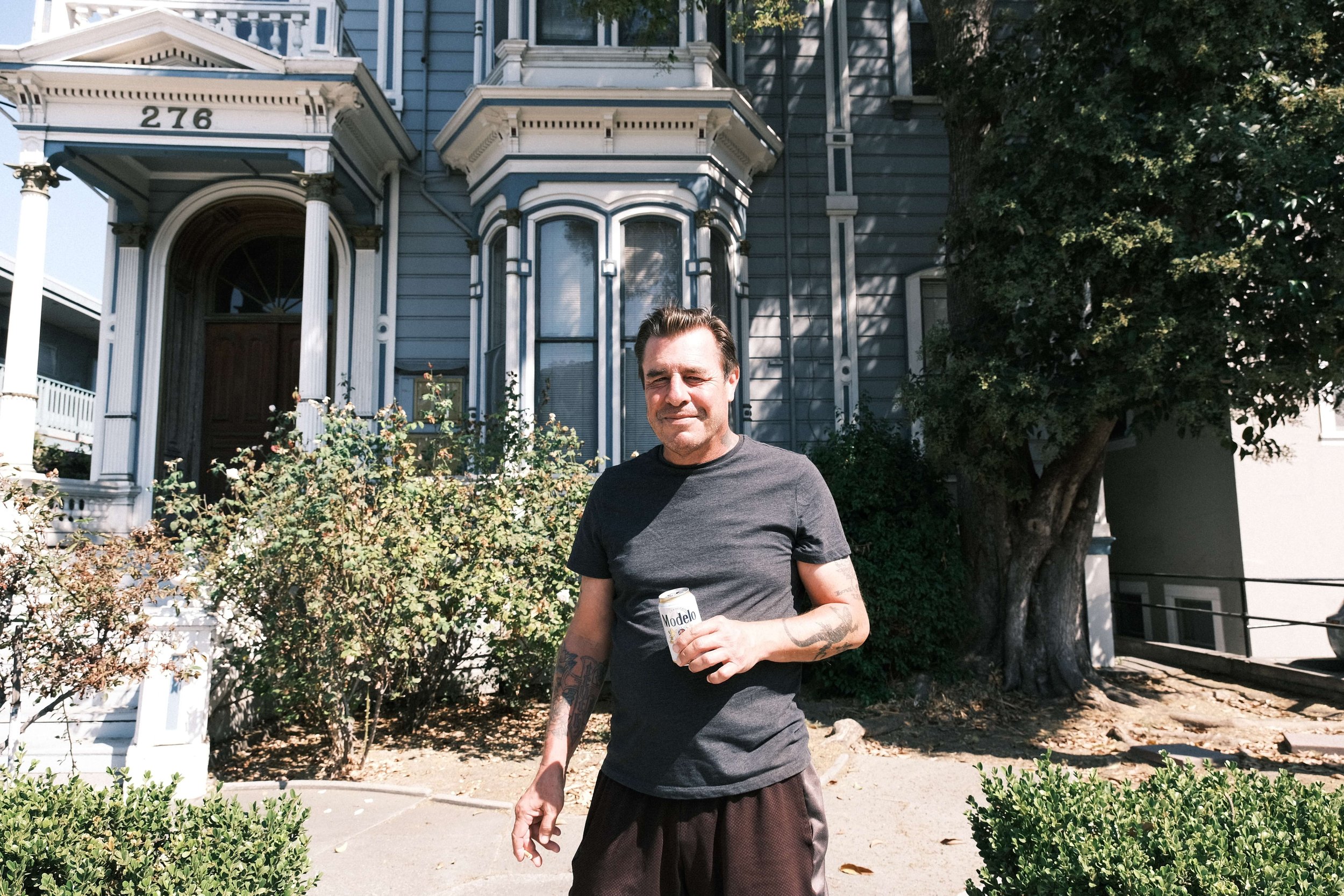 Man standing outside in front of a blue Victorian house, holding a can of Modelo beer, with bushes and a tree nearby.
