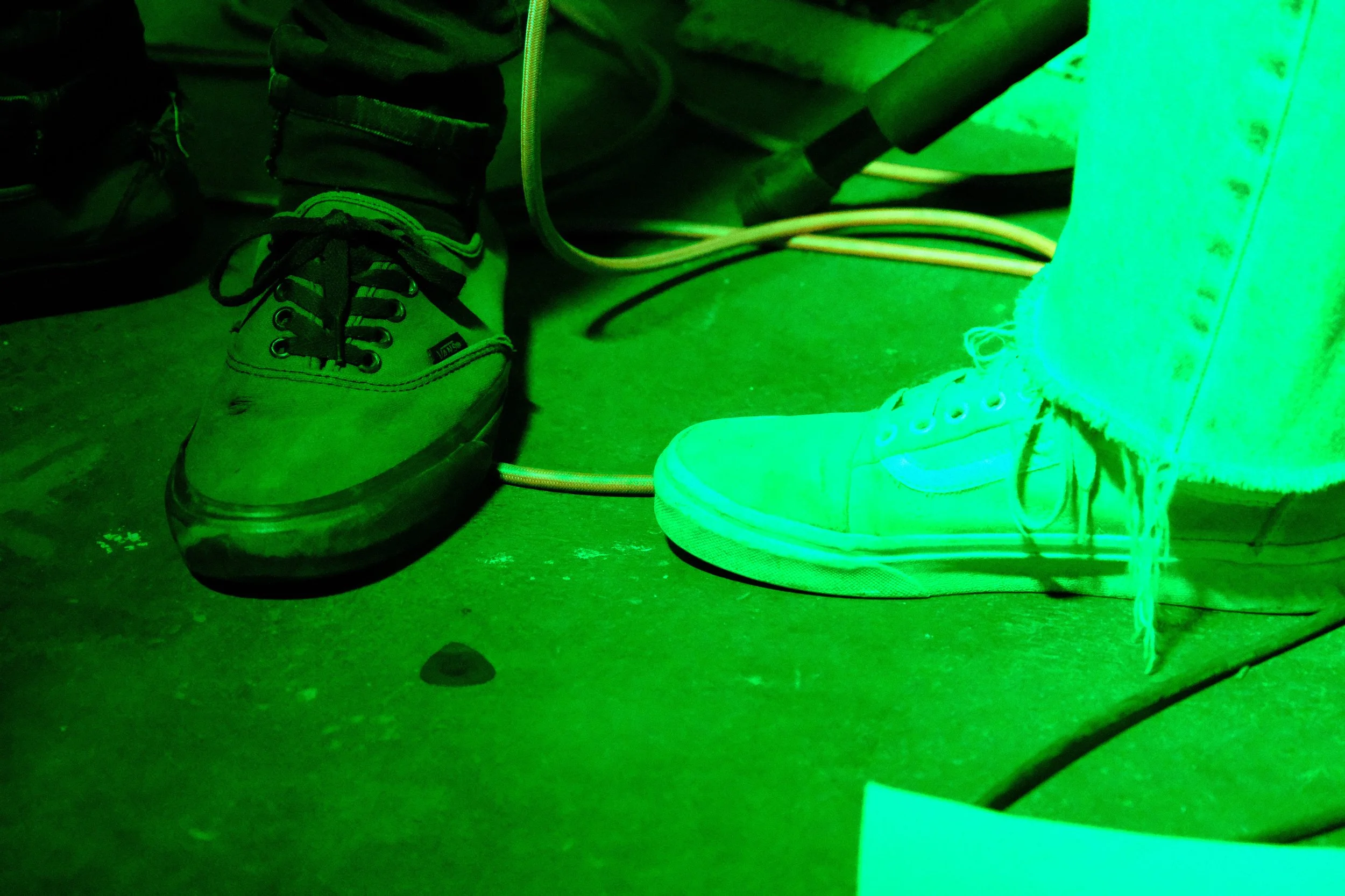 Close-up of two different sneakers on a floor, with wires and cables in the background, illuminated by green light.