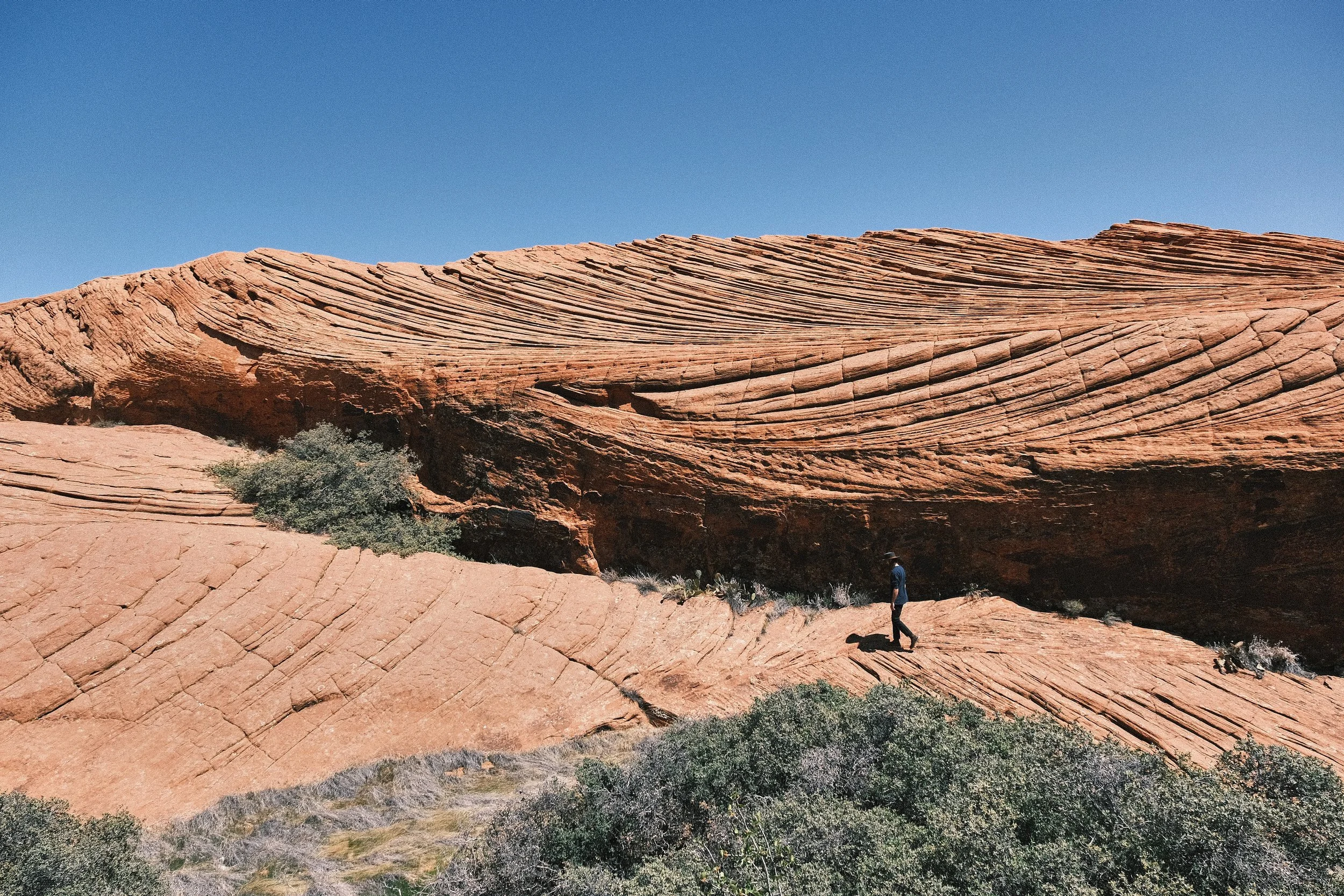 A person hiking on a large, layered red sandstone rock formation in a desert landscape under a clear blue sky.