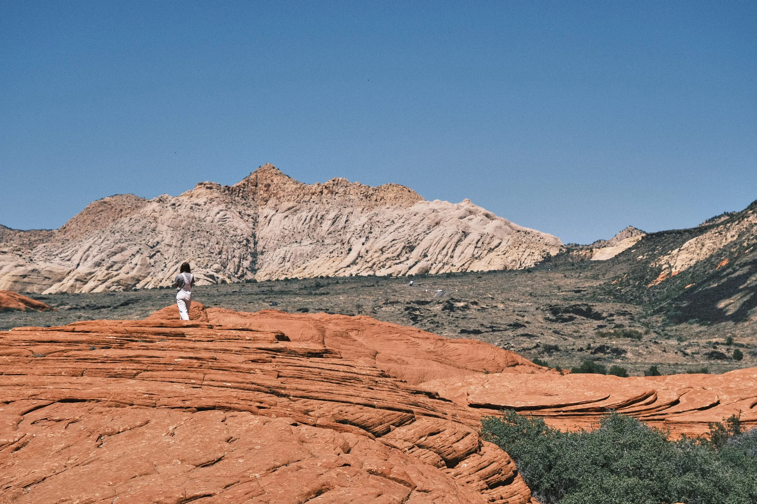 A woman in white standing on reddish rock formations in a desert landscape with large striped mountain in the background under a clear blue sky.