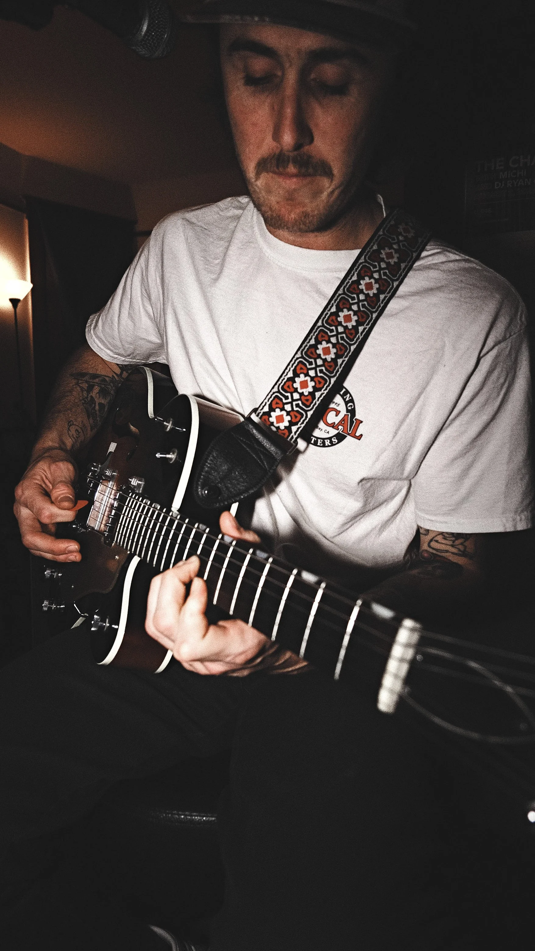 A man playing an acoustic guitar in a dimly lit room, wearing a white T-shirt and a baseball cap.