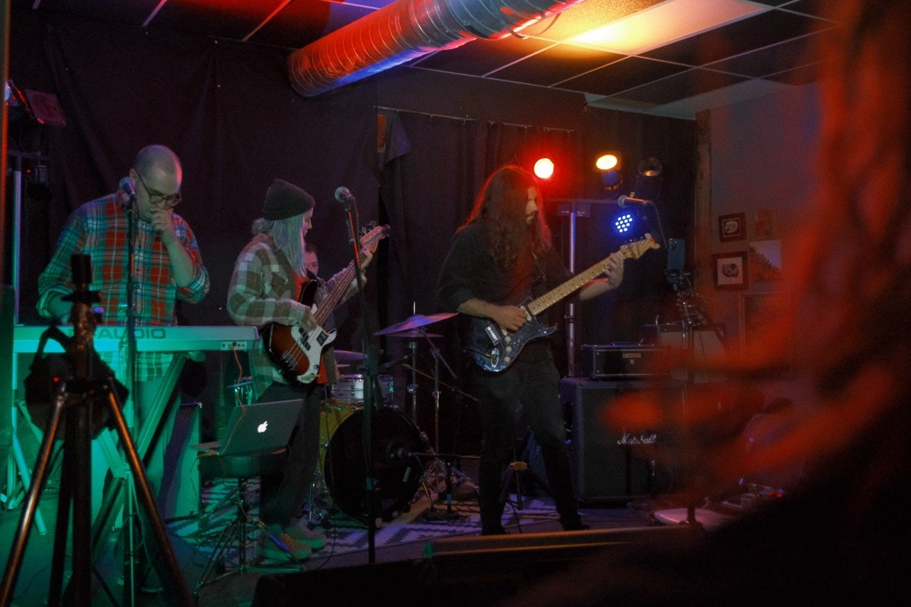 Band performing on stage with members playing keyboard, bass guitar, electric guitar, and drums, in a dimly lit venue with colorful stage lighting.