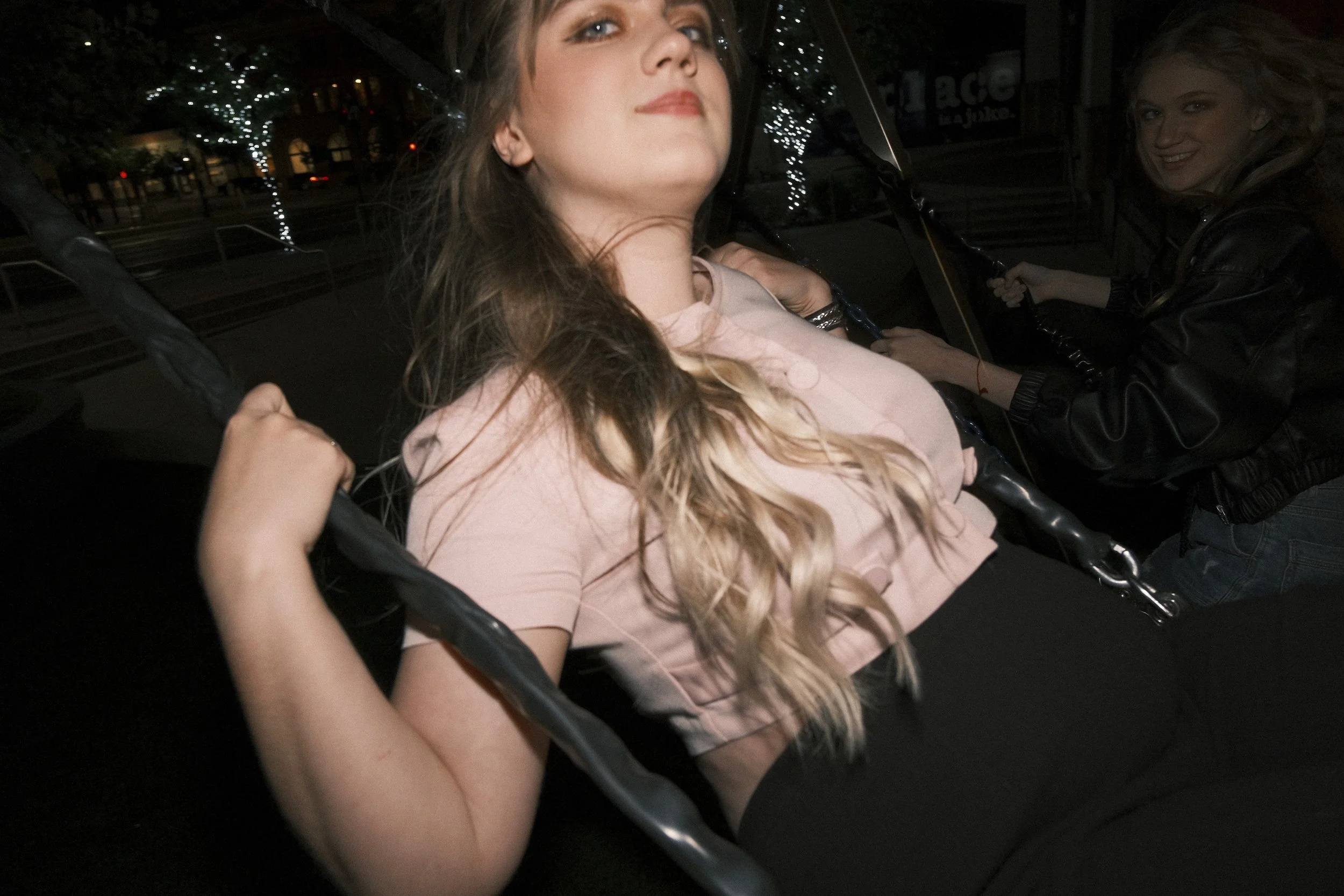 A woman sitting on a swing, looking confident, with another woman in the background holding a swing chain, on a night street with illuminated trees.