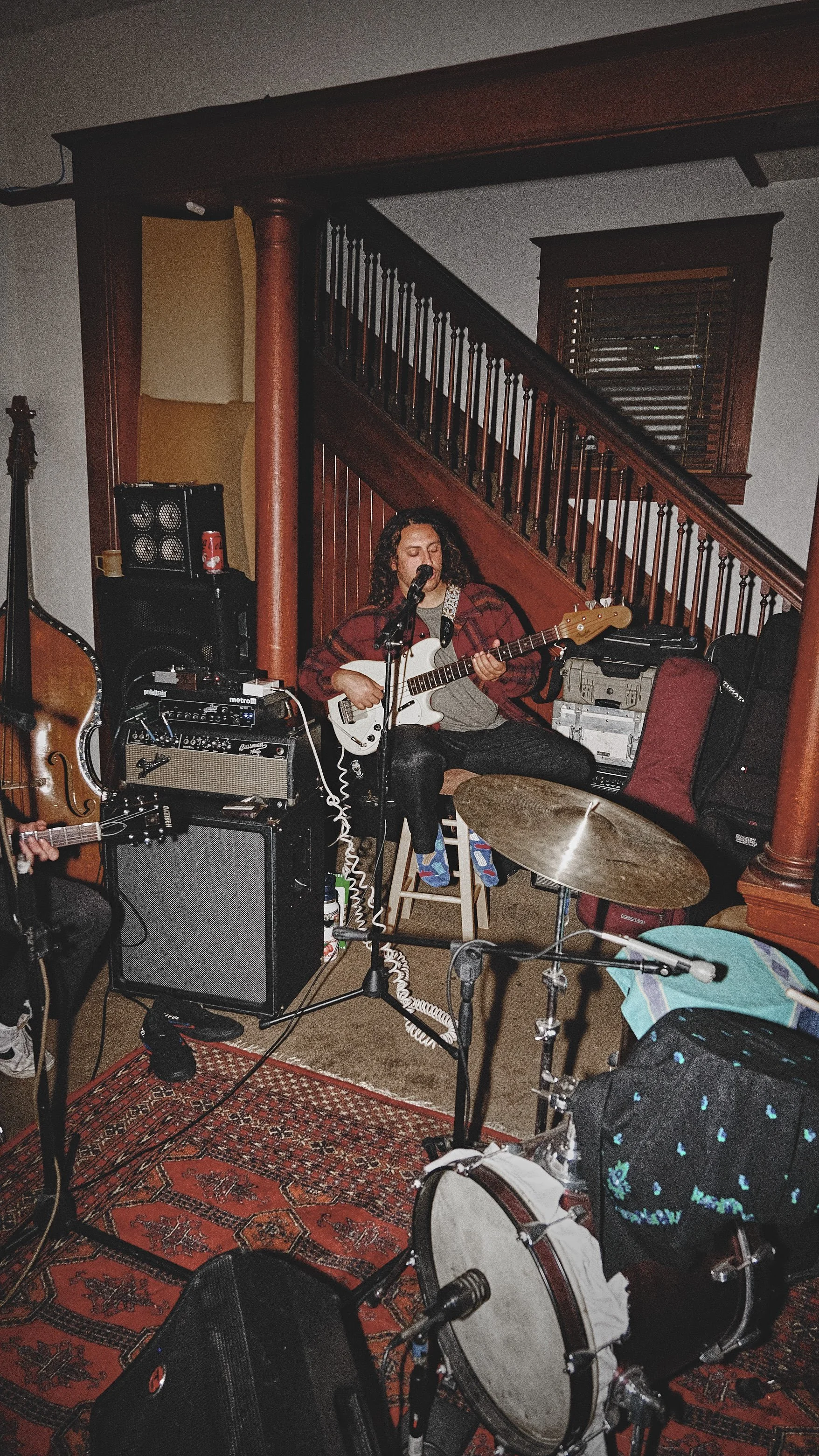 A person with curly hair playing electric guitar and singing into a microphone in a cozy, dimly lit room with musical equipment and a staircase in the background.