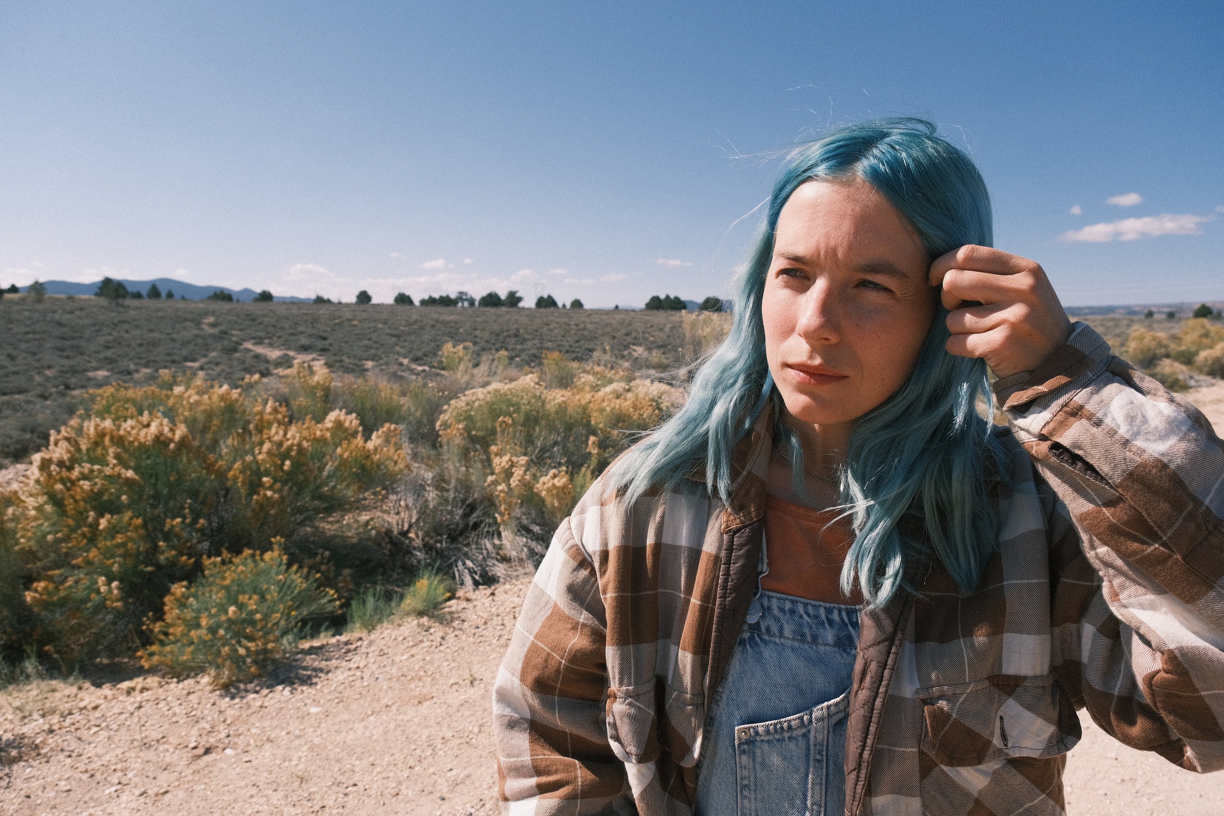 A young woman with blue hair looks to the side in a desert landscape with bushes and mountains.