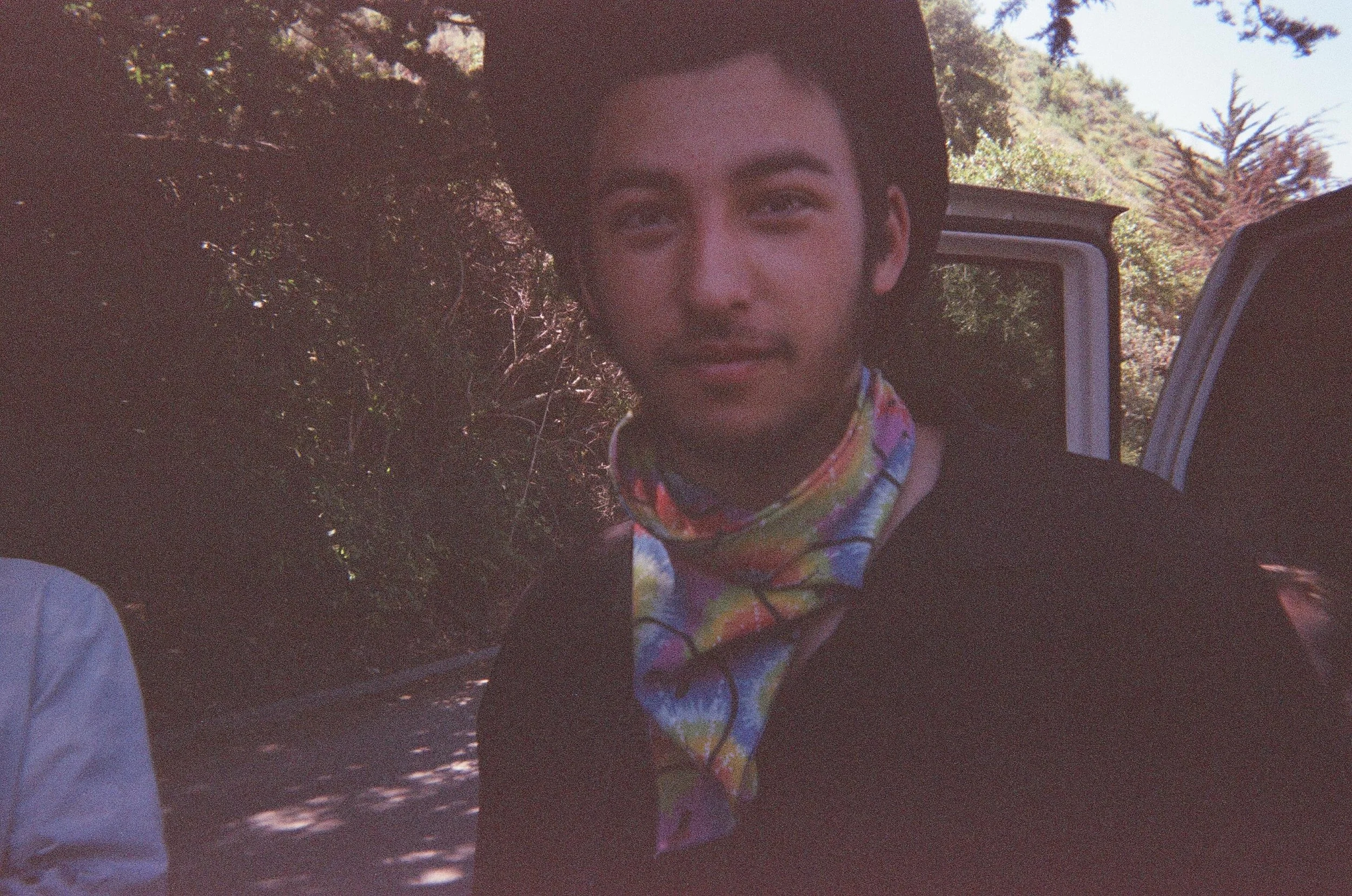 A young man with short dark hair and a facial tattoo, wearing a colorful bandana around his neck, standing outdoors near a vehicle with trees and mountains in the background.