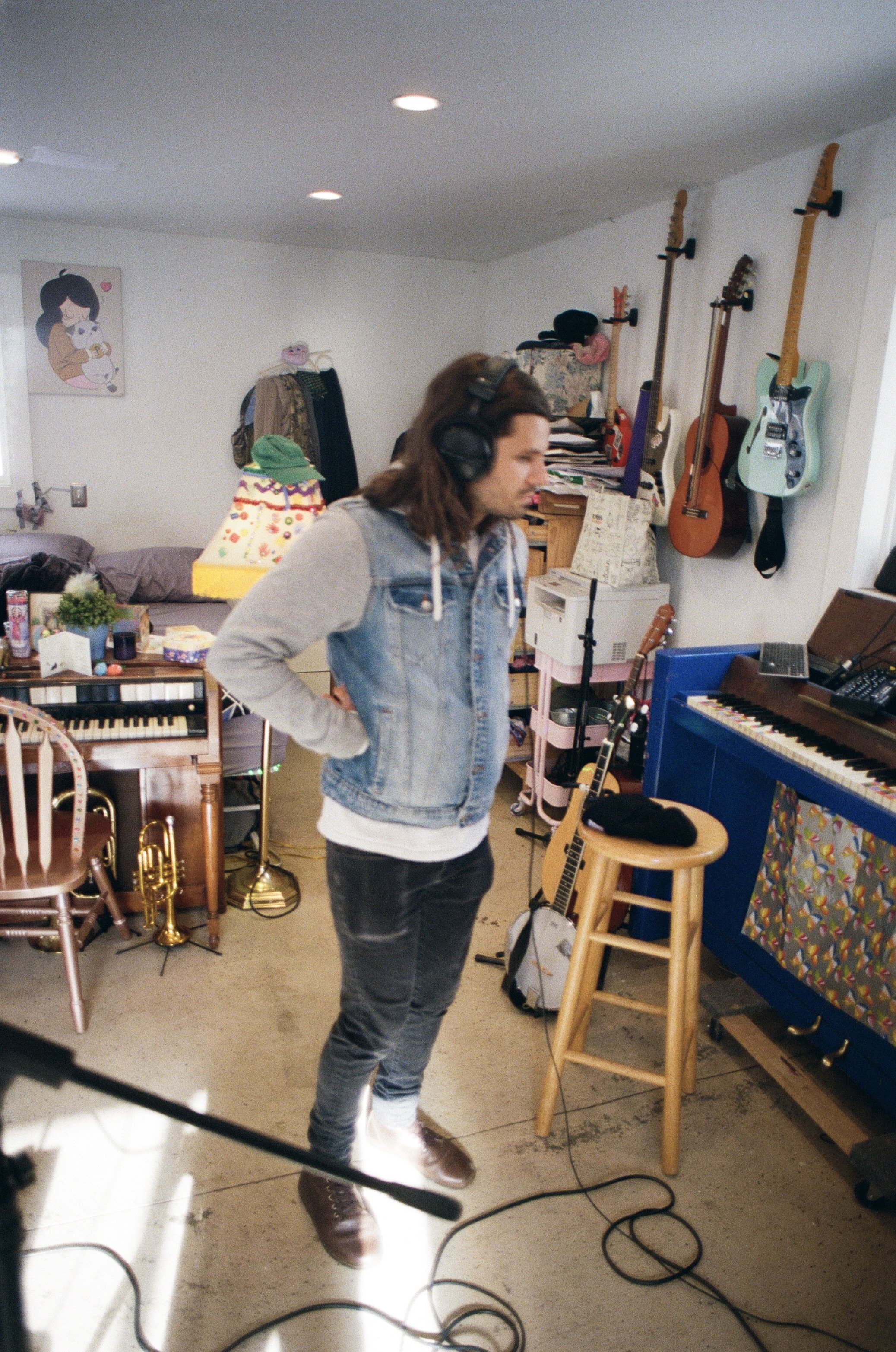 A person with long hair wearing headphones, a denim vest, and dark jeans, standing with hands on hips in a music or studio room filled with musical instruments including guitars, a keyboard, and a trumpet.