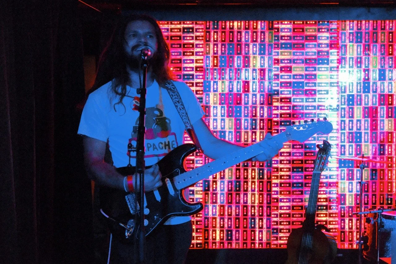 A man with long hair holding a guitar, standing in front of a colorful neon background with cassette tape patterns, on a stage with a microphone.