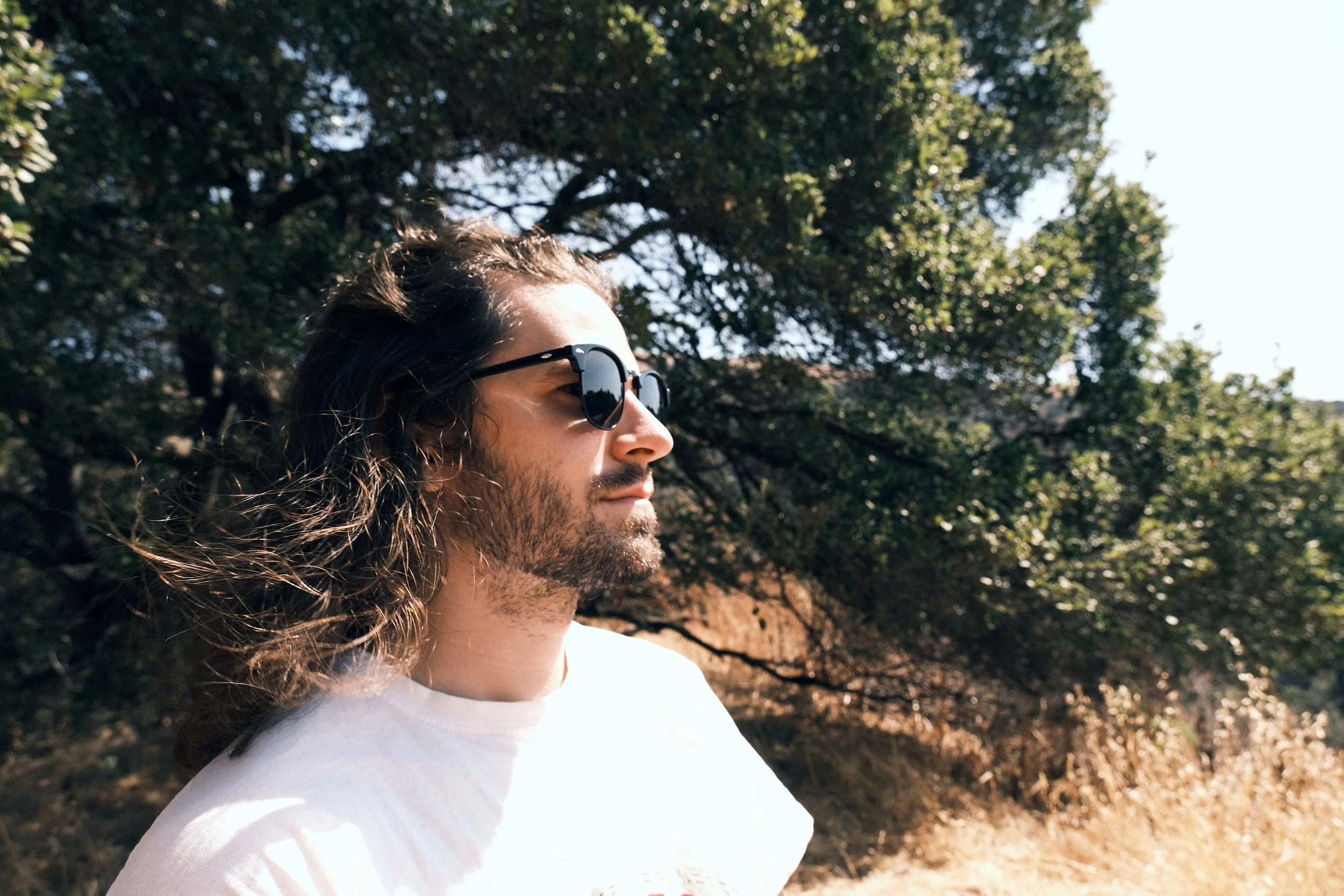 A man with long hair and a beard wearing sunglasses outdoors, standing in front of trees and a sunny sky.