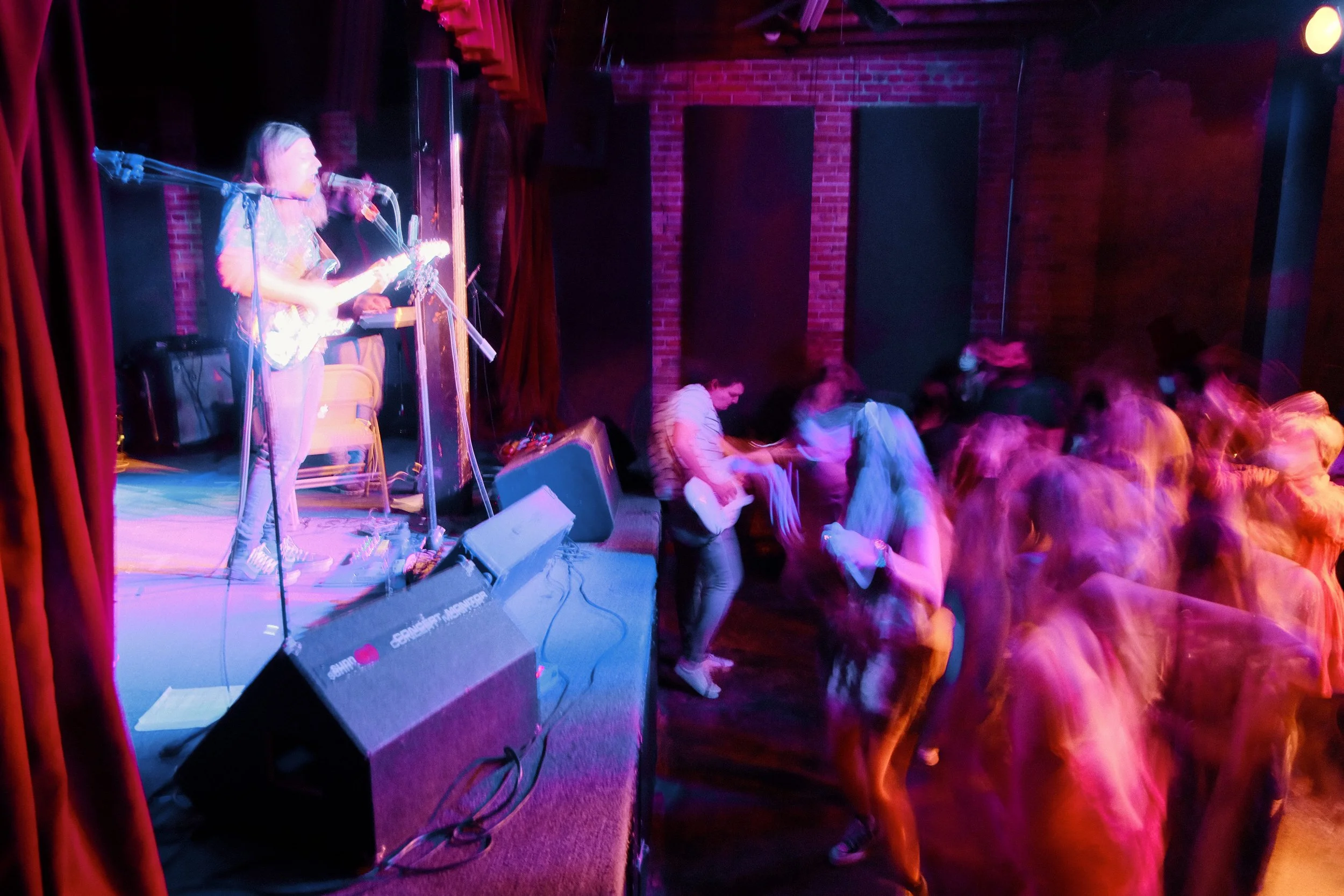 Musician performing on stage with an electric guitar at a dimly lit venue, audience dancing near the stage, expectant of live music.