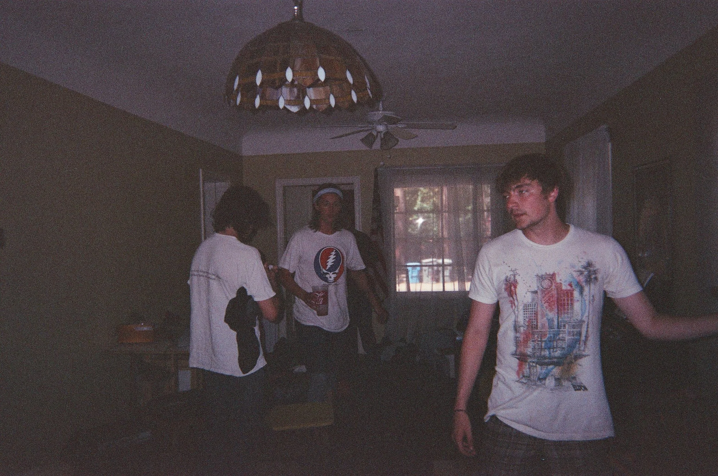 Three young men in a dimly lit room, with one in the foreground wearing a graphic T-shirt and two in the background, one with a white T-shirt and the other with a Bo Diddley shirt, standing near a window with curtains, under a ceiling fan and a vinta