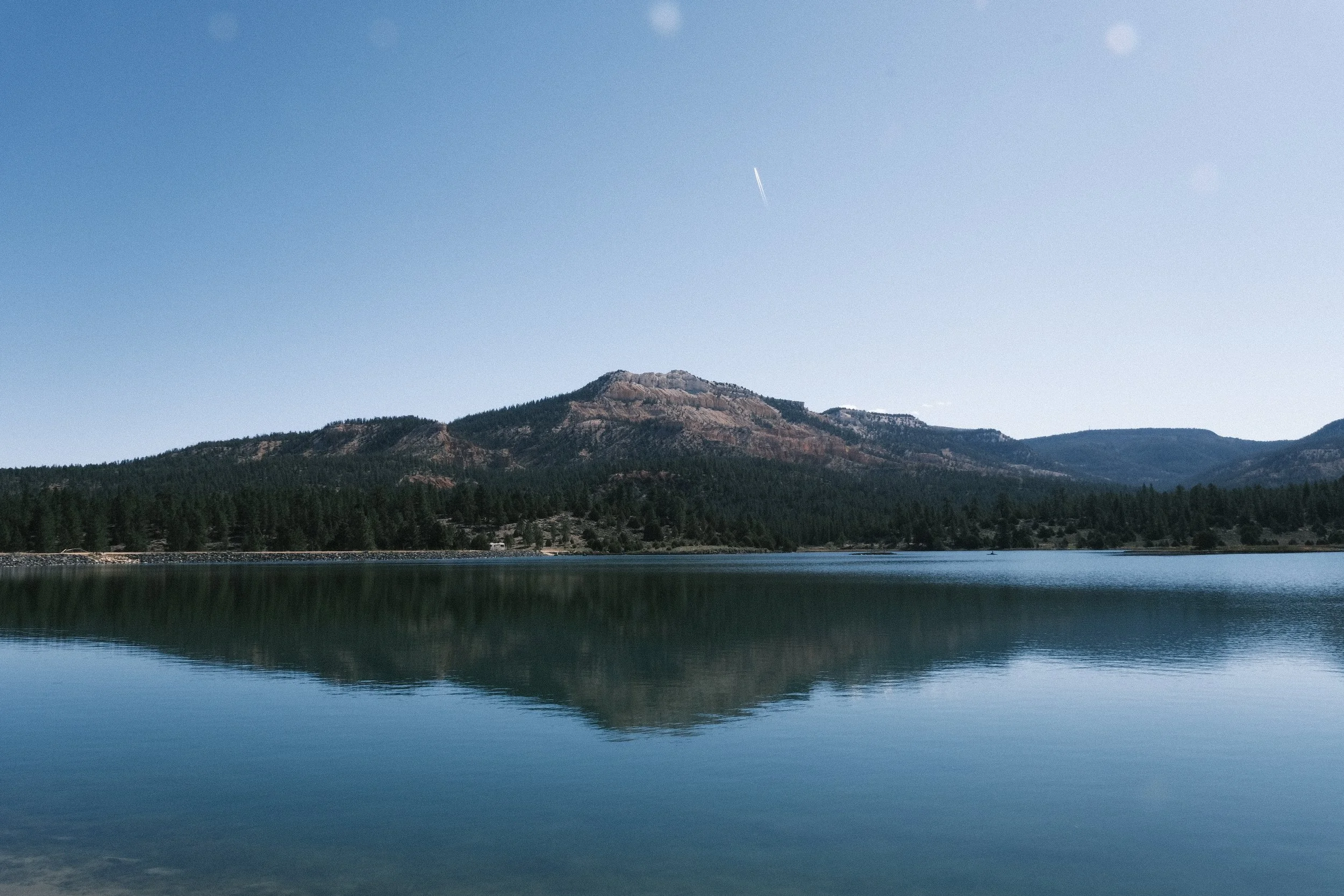 A calm lake reflects a forested mountain and blue sky with a faint contrail.