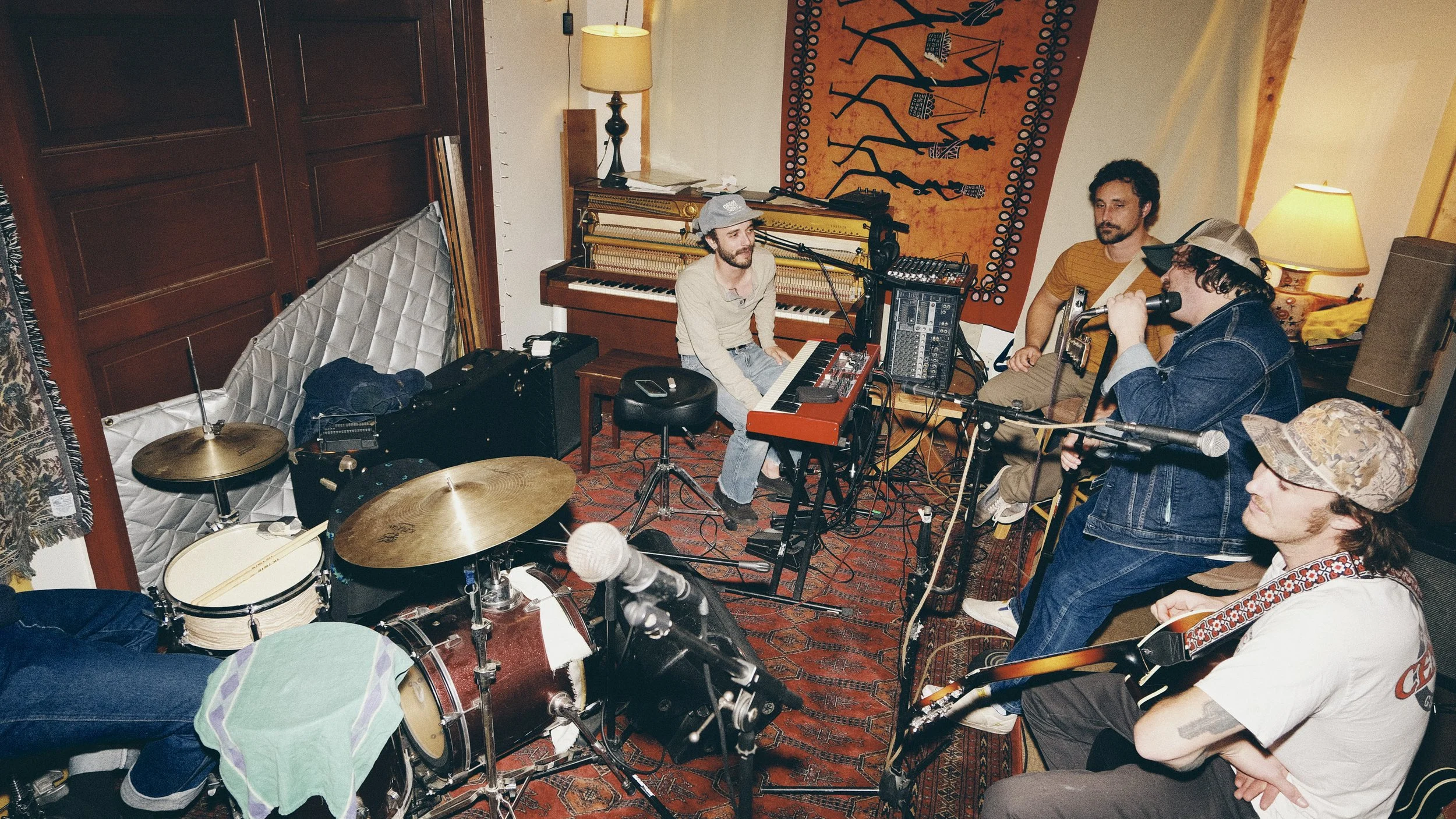 Four musicians in a cozy, lived-in studio, with one singing into a microphone, one playing the guitar, one on keyboard, and the fourth on drums, surrounded by various musical equipment, lamps, and a colorful tapestry on the wall.