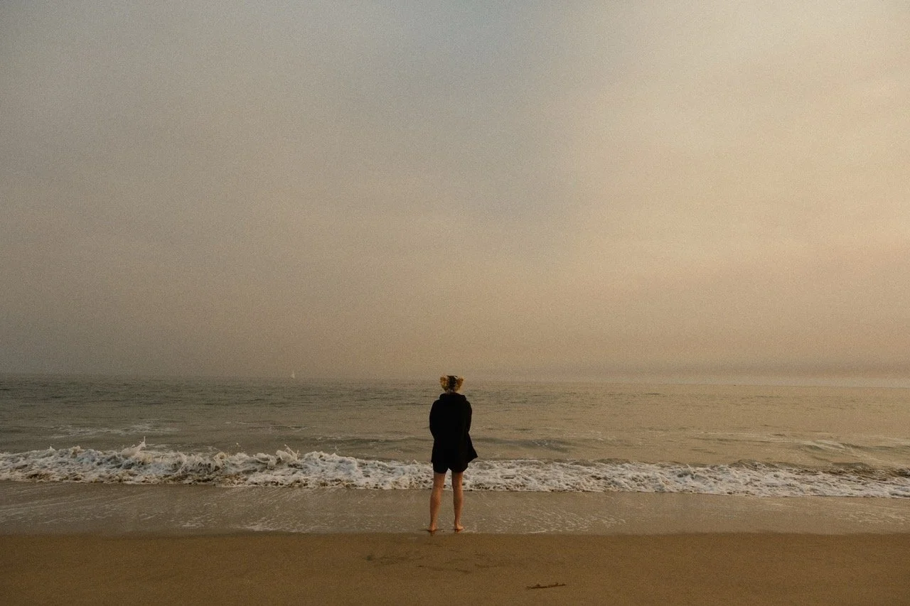 A person standing on the beach facing the ocean during sunset, wearing a black hoodie and shorts.
