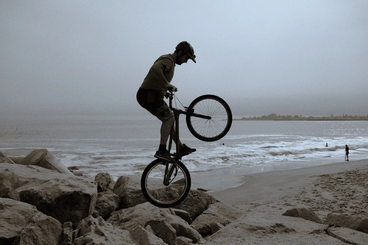 Person in a helmet balancing on a unicycle on rocks near the beach during overcast weather.