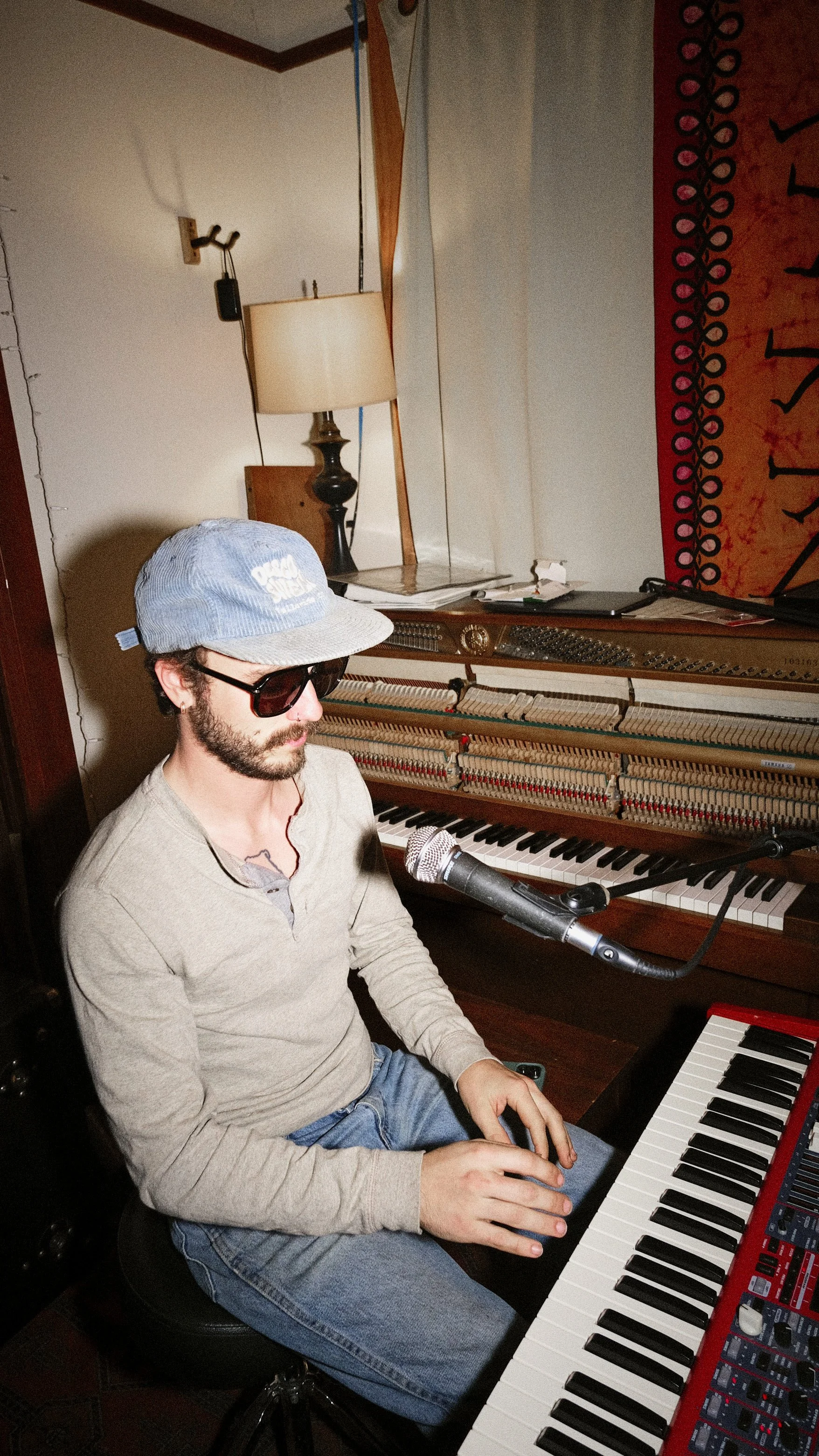 A man wearing sunglasses, a gray cap, a beige long-sleeve shirt, and jeans is sitting at a keyboard piano with a microphone positioned in front of him. He is in a room with a lamp, a wall hanging, and an open grand piano behind him.