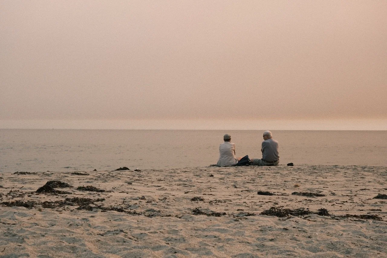 An elderly man and woman sitting on a sandy beach facing the ocean at sunset or sunrise.