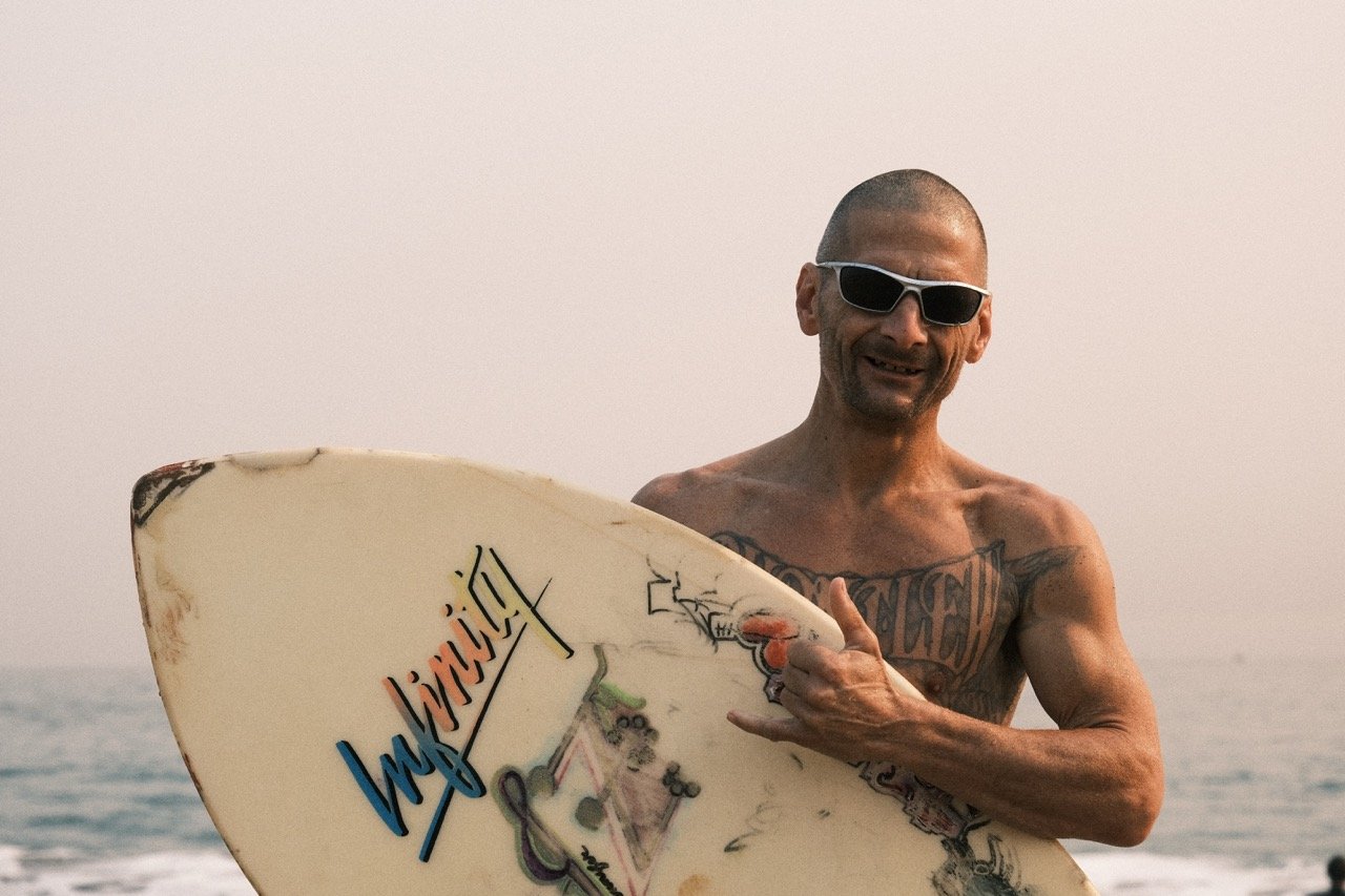 A shirtless man with sunglasses holding a surfboard at the beach, smiling.
