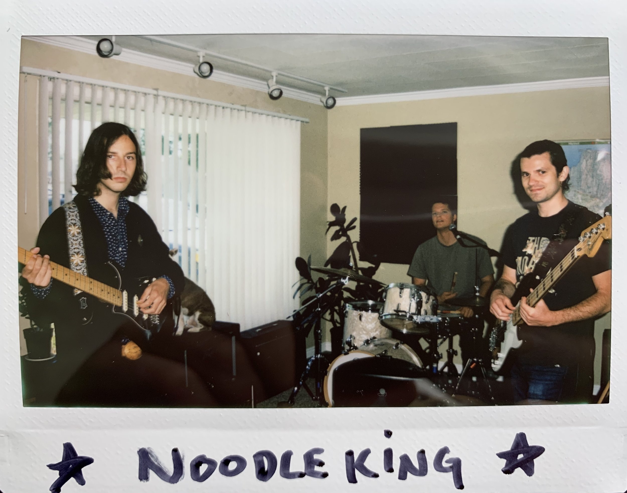 Three young men in a living room, playing musical instruments. One on the left with long dark hair and a guitar, one in the center behind a drum set, and one on the right with short dark hair holding a guitar. There are vertical blinds on the window 