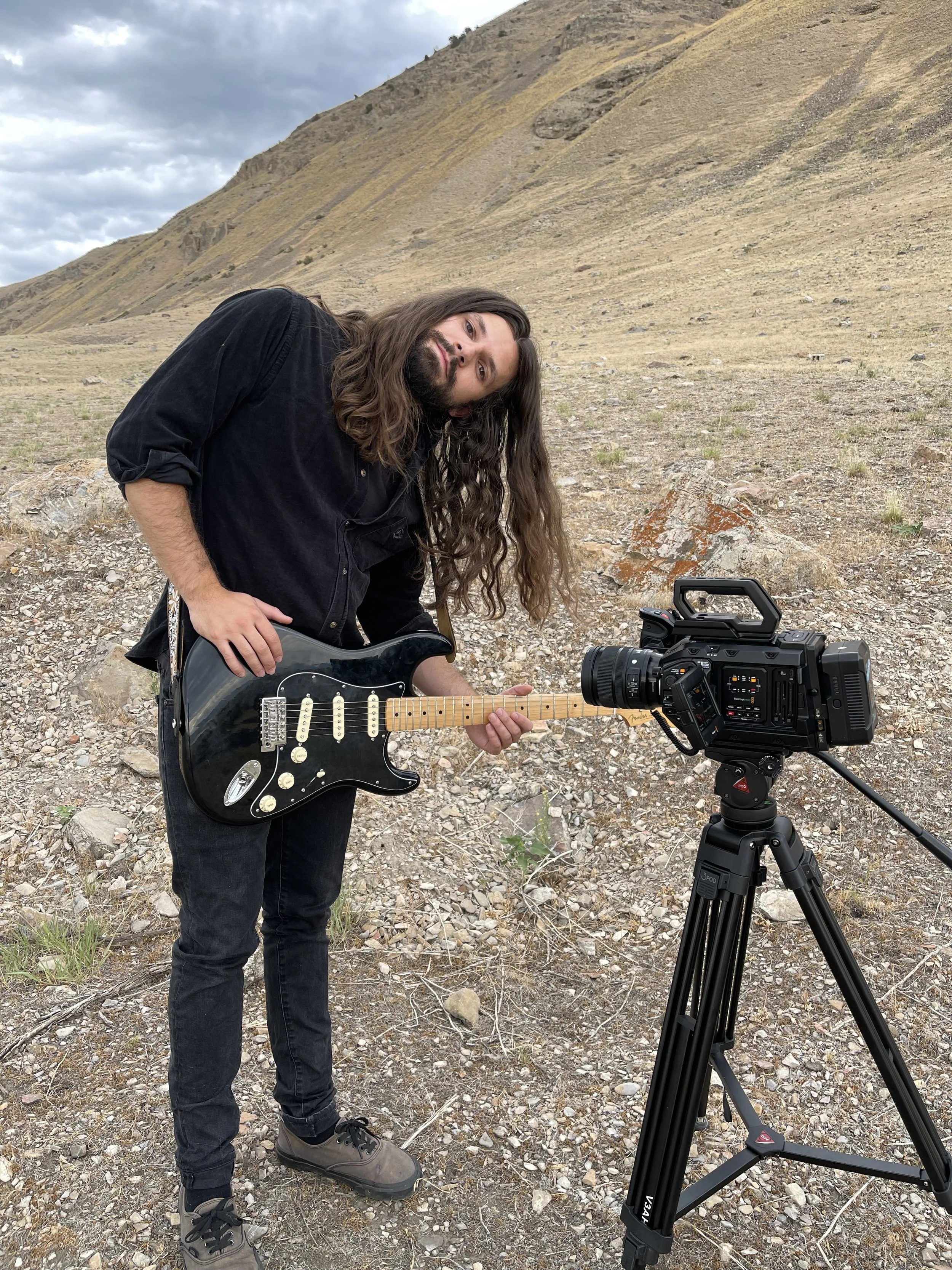 A person with long hair and a beard wearing a black shirt and jeans, holding a black electric guitar, stands outdoors in a rocky, mountainous landscape with a camera on a tripod in front of him.