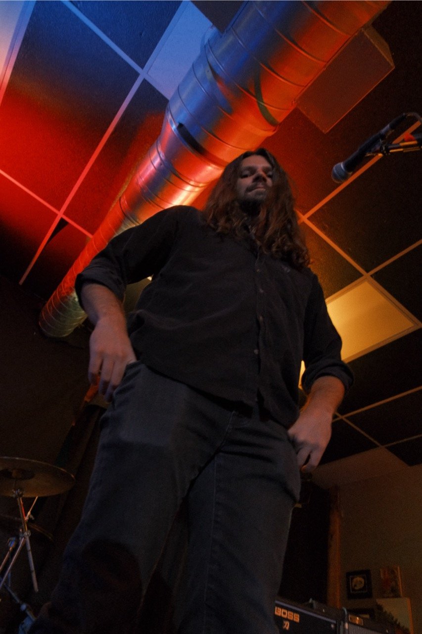 A man with long brown hair in black clothing standing on stage near a microphone with a drum set in the background, illuminated by red and orange stage lighting.