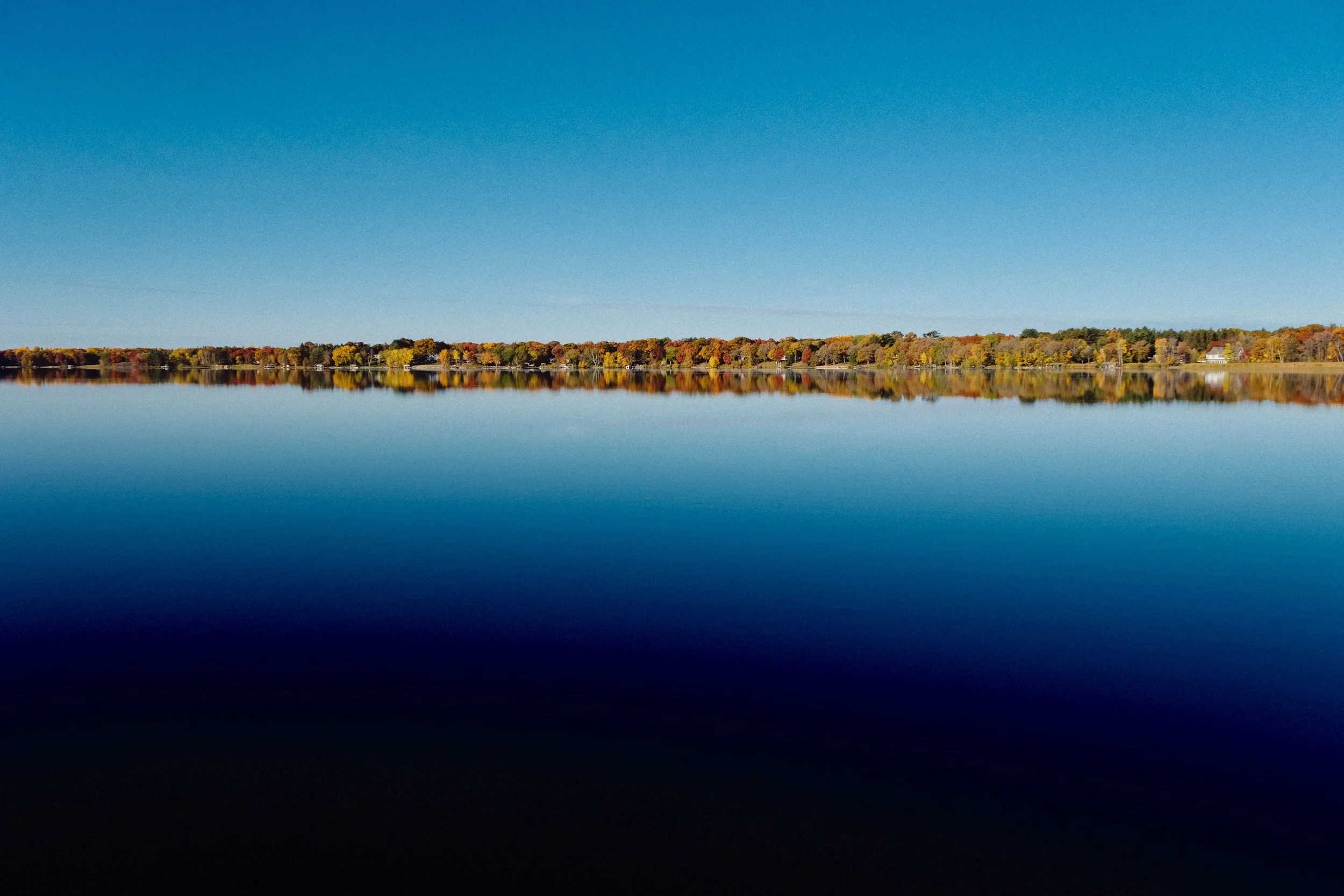 Calm lake reflecting a line of trees with autumn foliage under a clear blue sky.