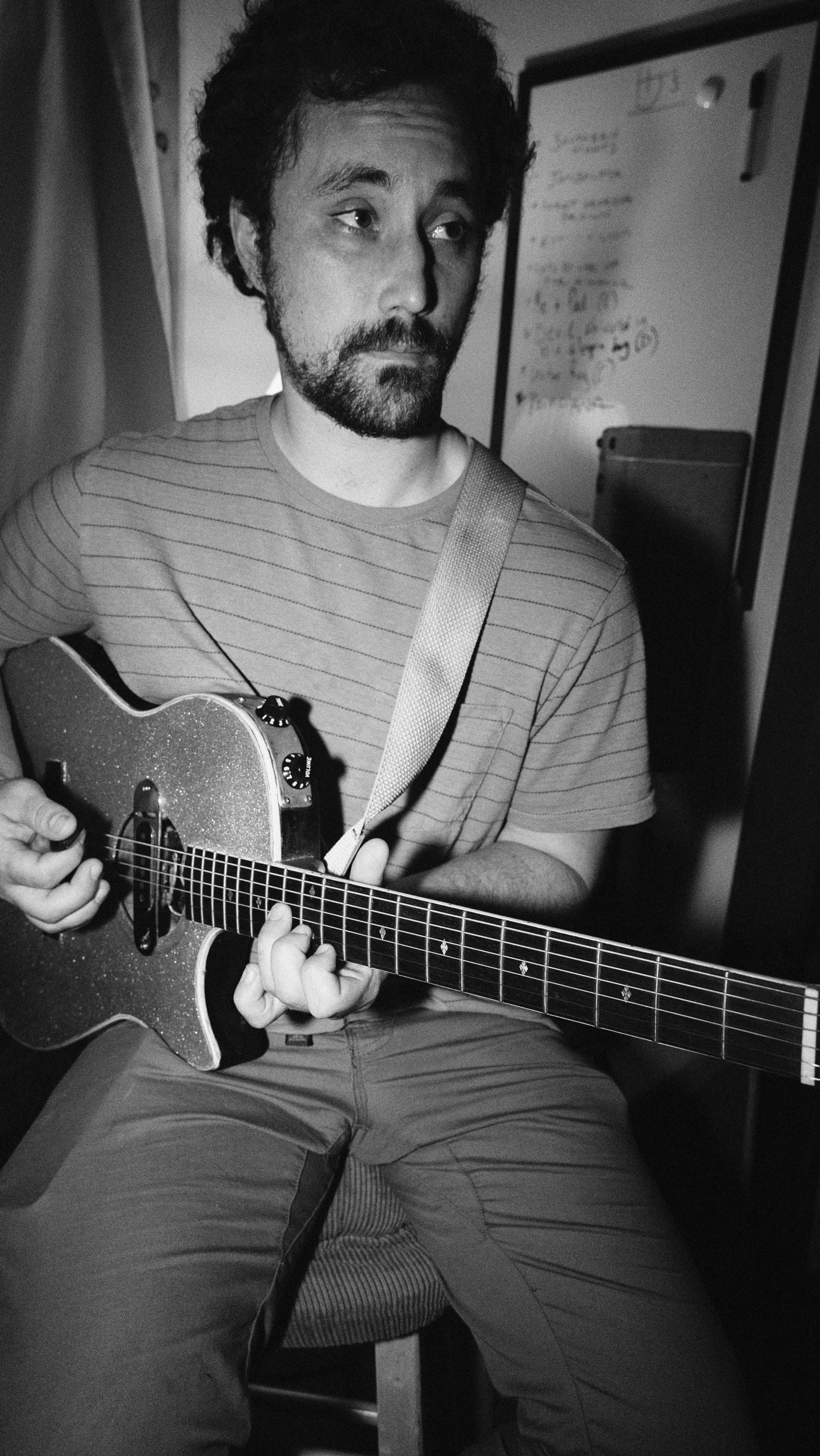 Black and white photo of a man with dark, curly hair and a beard playing an acoustic guitar, wearing a striped t-shirt, sitting on a stool in front of a whiteboard.