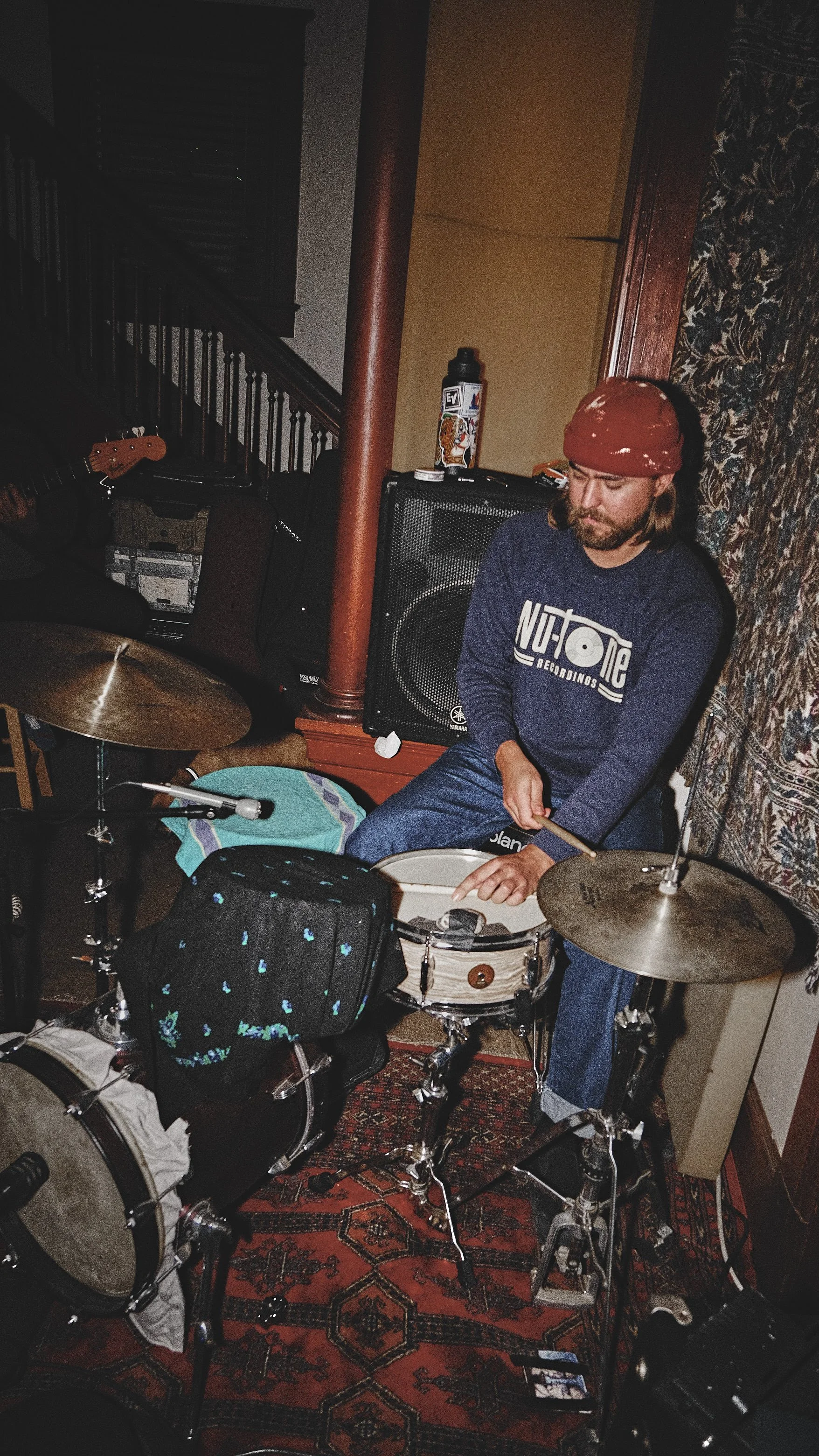 A man with long hair, beard, wearing a red bandana and blue sweatshirt sitting while playing drums in a cozy room with a rug and musical equipment.