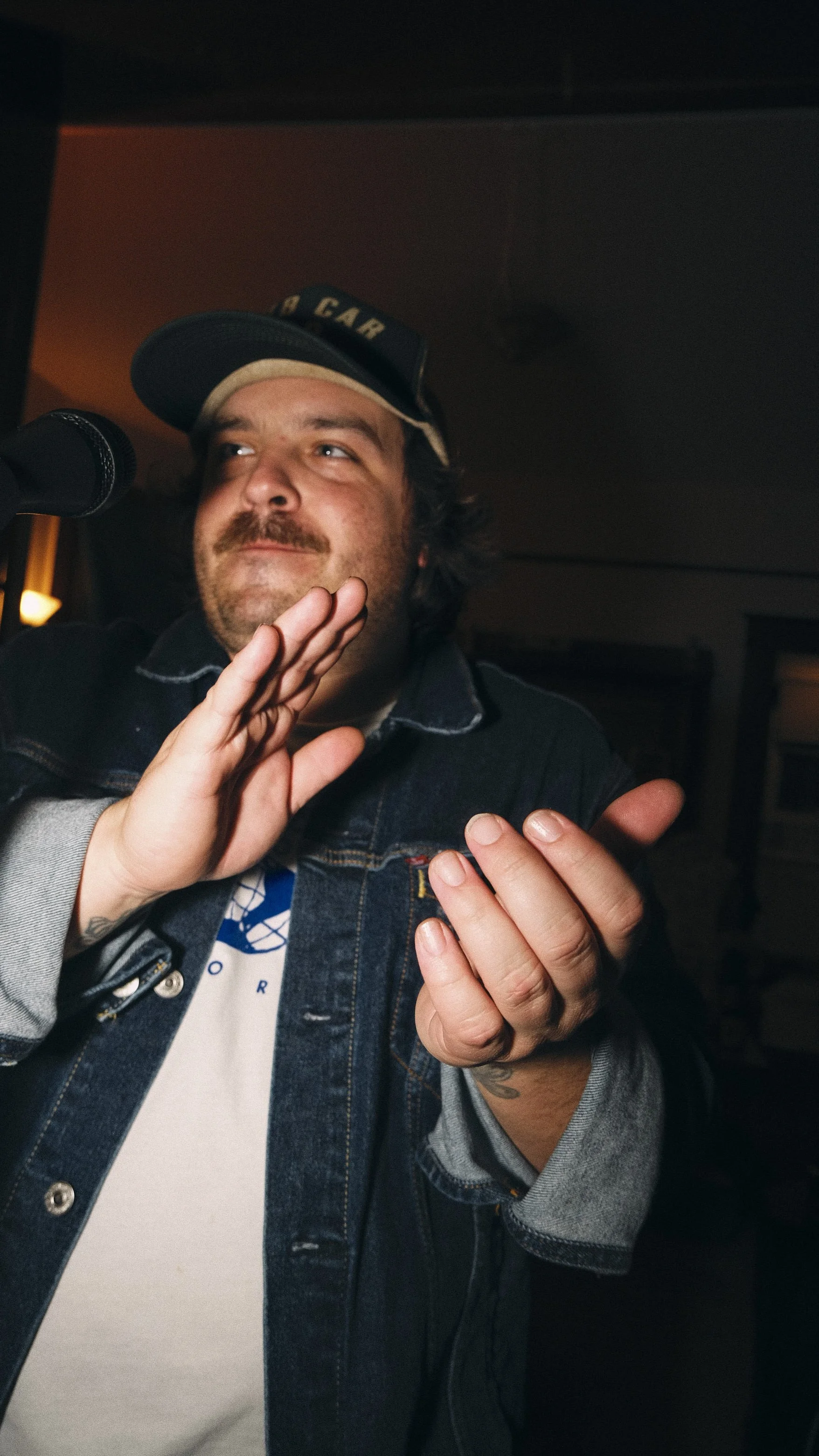 Man with beard and mustache, wearing a black denim jacket, beige hat with text, standing near microphone, gesturing with hand, in indoor setting.