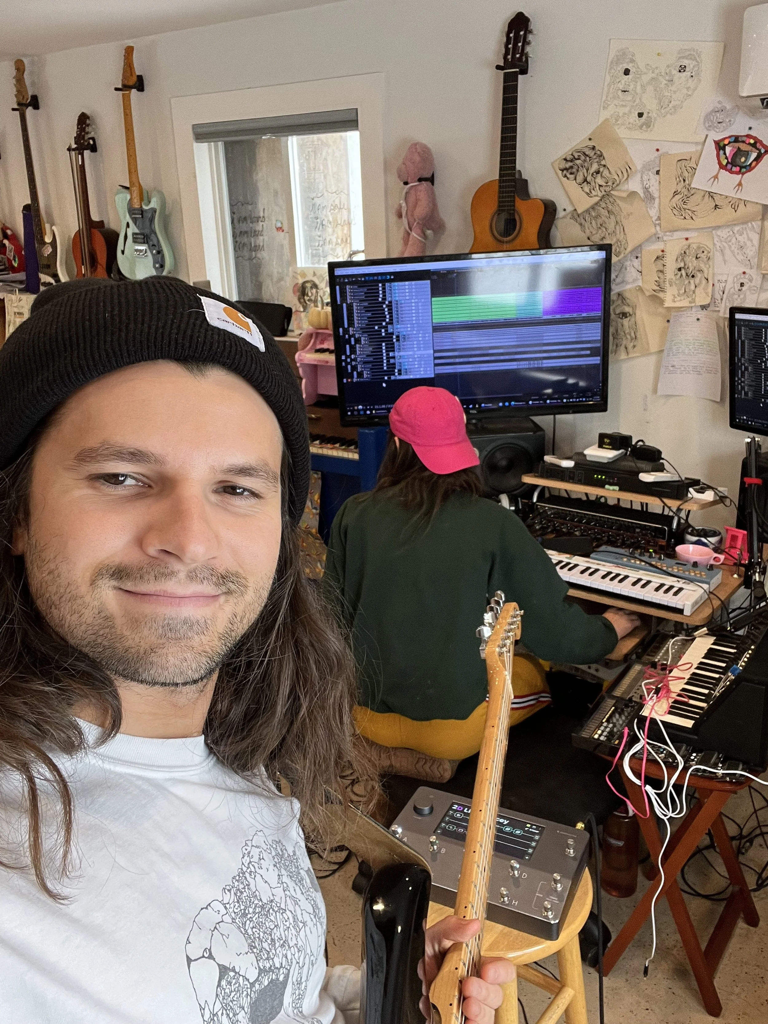 A man with long hair, a beard, and a black beanie is taking a selfie in a music studio. Behind him, a person with a pink cap is working at a keyboard with various musical equipment. The room has guitars hanging on the wall, sketches, and artwork pinn