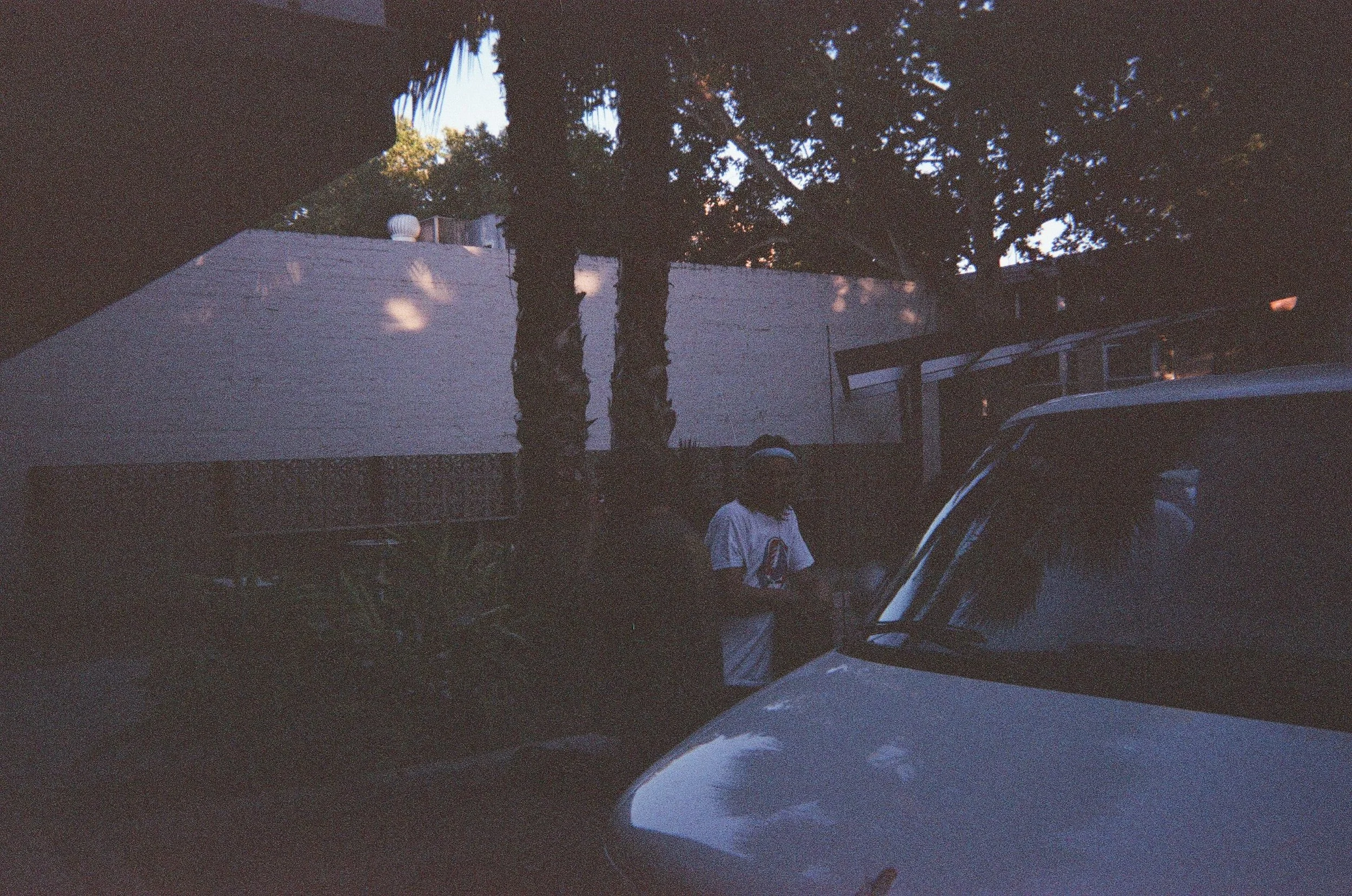 A person standing next to a white vehicle, with trees and a white building in the background, during what appears to be overcast or low-light conditions.