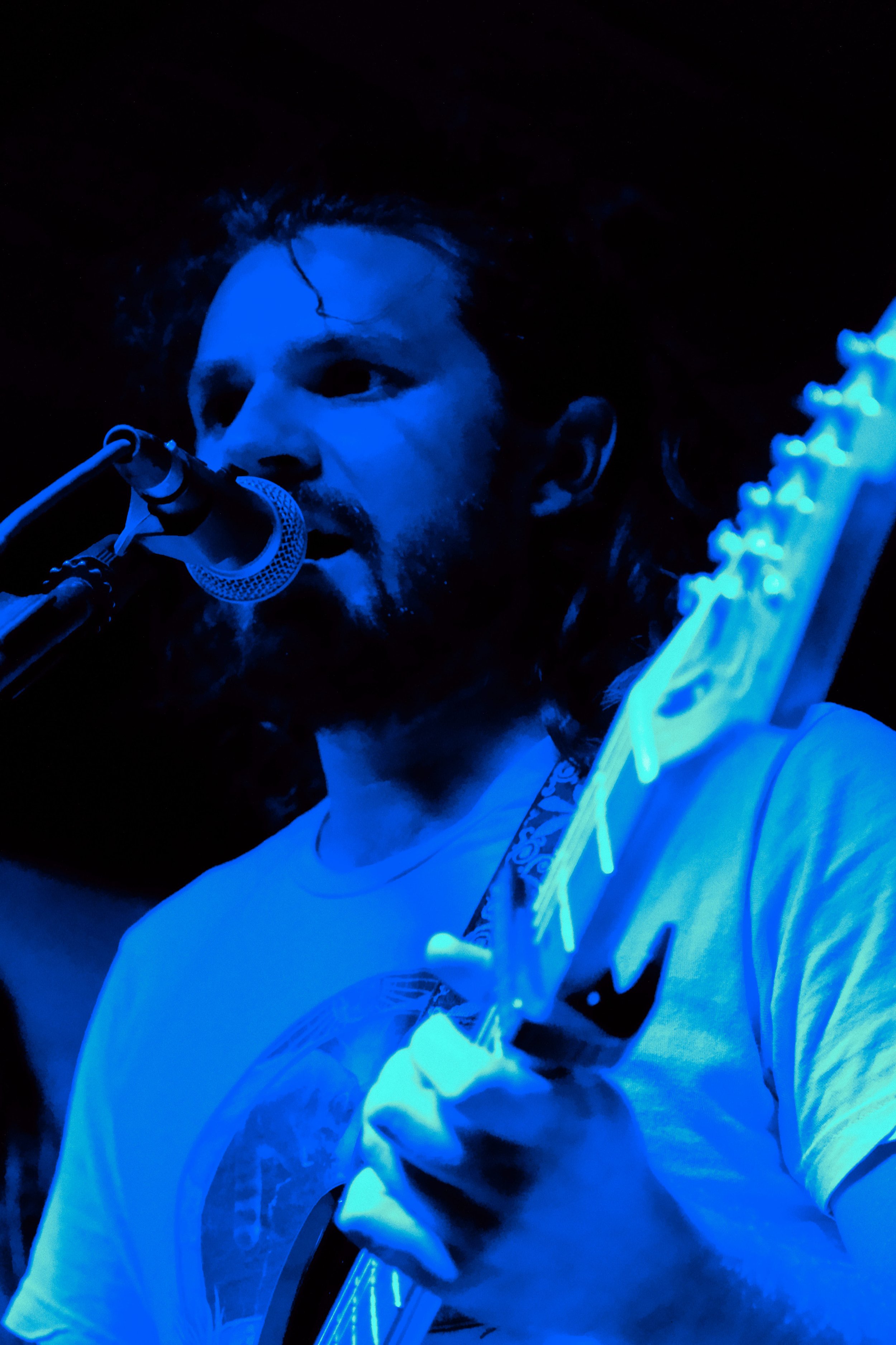 Musician with long hair and beard singing into a microphone while playing an electric guitar, illuminated by blue lighting.