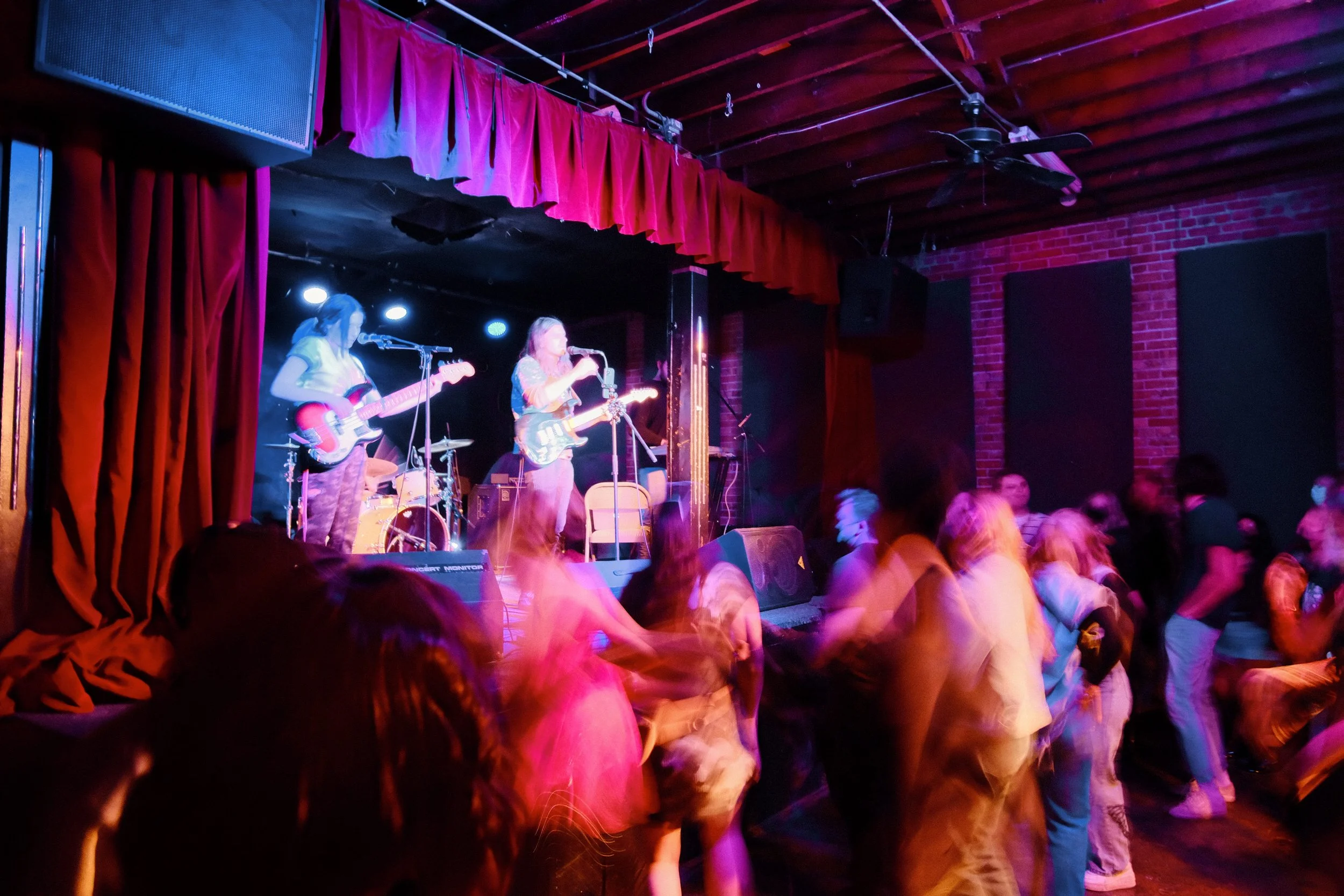 A live music performance at a nightclub with two female guitarists on stage, colorful lighting, red curtains, and people dancing in front.