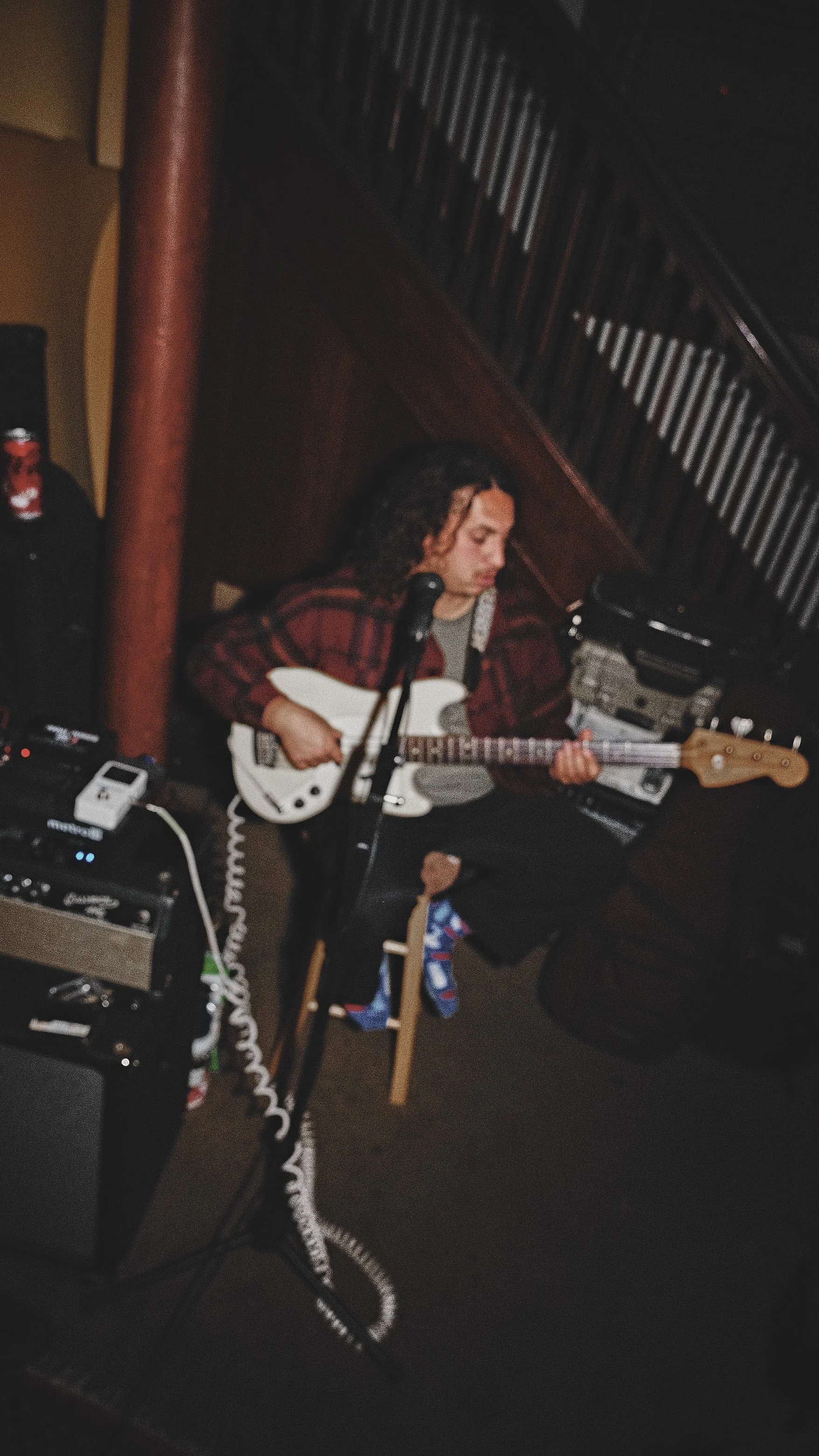 A person sitting on a small stool playing an electric guitar, surrounded by music equipment in a dimly lit room.