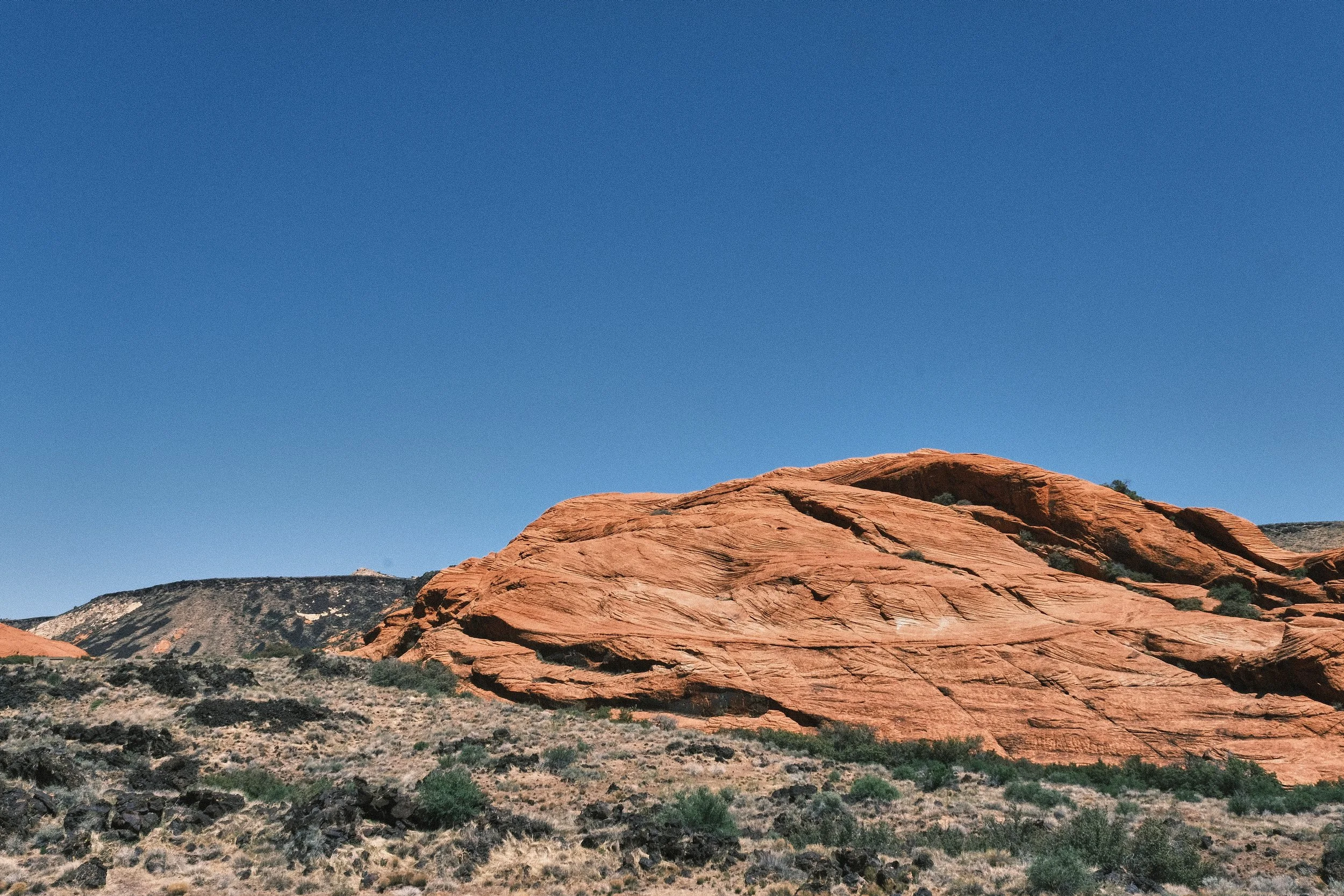 Red sandstone rock formation in a desert landscape with sparse vegetation under a clear blue sky.
