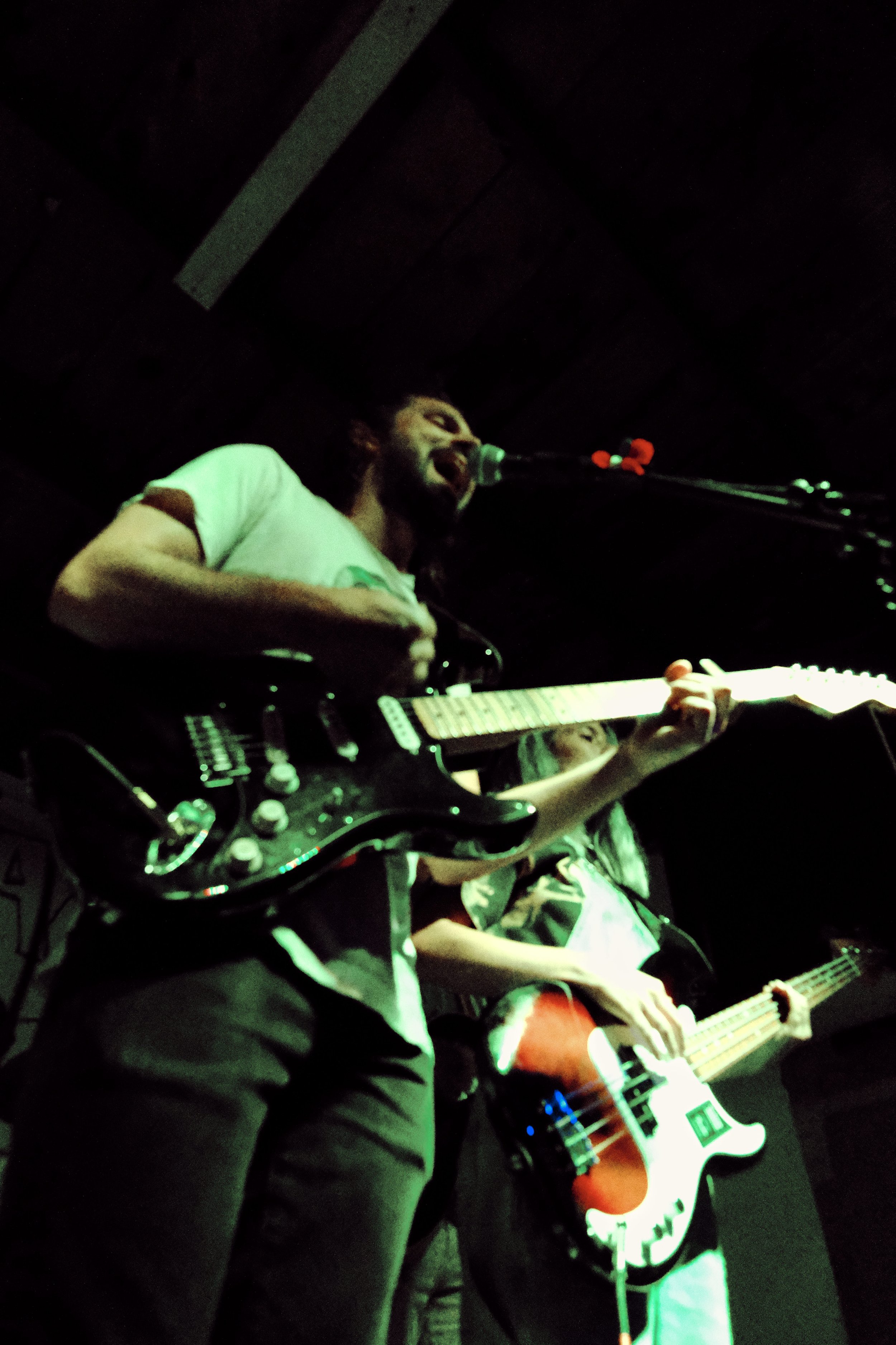 A musician singing into a microphone while playing an electric guitar on stage in a dimly lit setting.