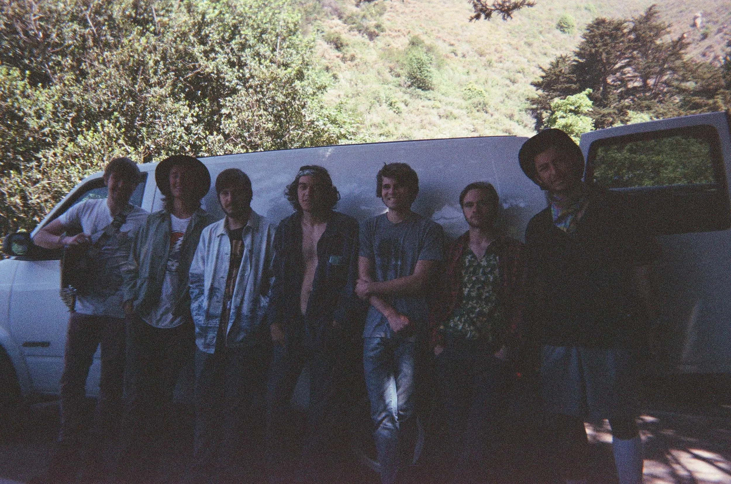 A group of seven people standing in front of a white vehicle, with a background of green trees and hills.
