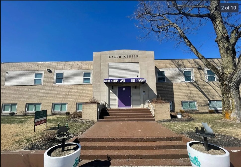 Exterior view of a building labeled as Labor Center, with steps leading up to a purple door, and a sign indicating 'Labor Center Lofts, 1125 S Walnut.' There are window planters with decorative bird statues, a large tree to the right, and a clear blu