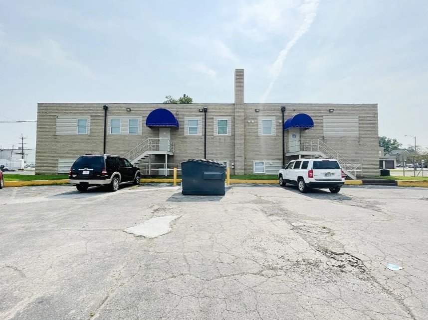 A two-story beige building with blue awnings over the windows and staircases, parked vehicles, a large trash bin, and a cracked asphalt parking lot.