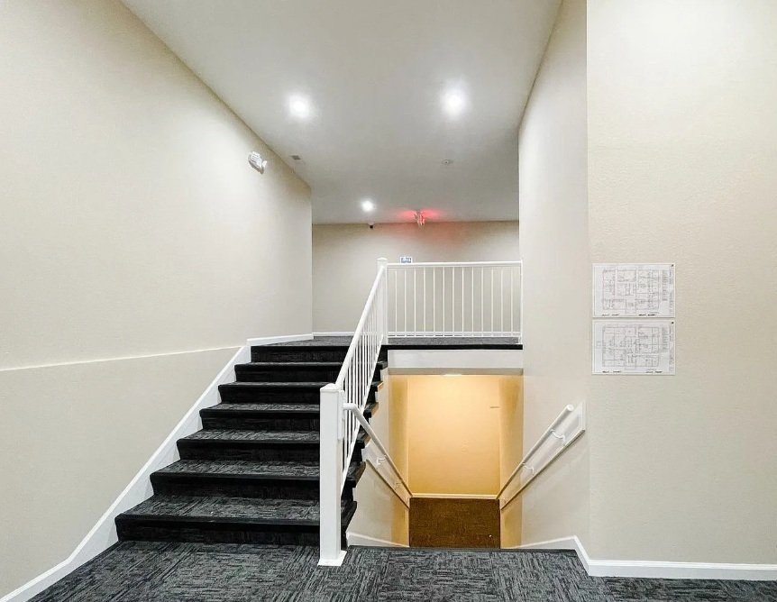 Indoor staircase with black carpeted steps, white railings, beige walls, and ceiling lights leading down to a lower level in a building.
