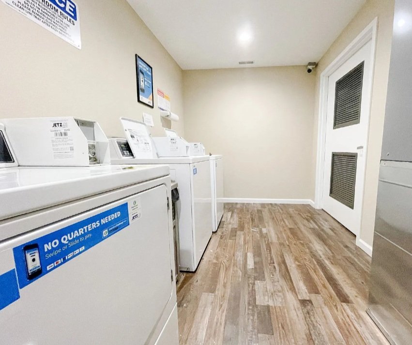 Laundry room with four washing machines and a dryer, wood flooring, beige walls, and a door with ventilation grilles.