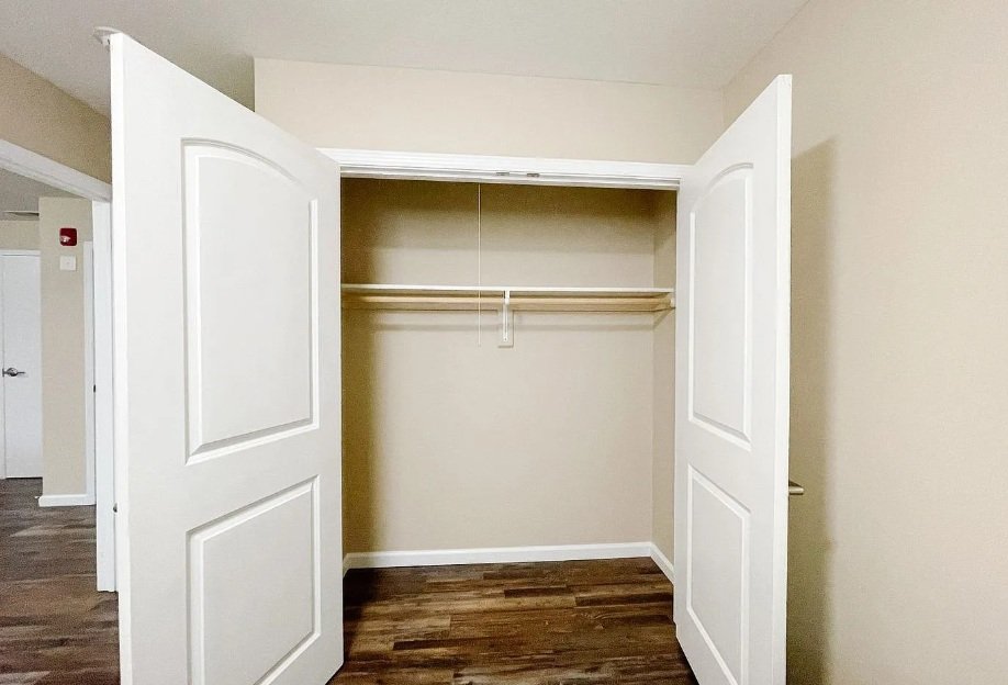 Empty closet with white double doors, beige walls, and hardwood floors.