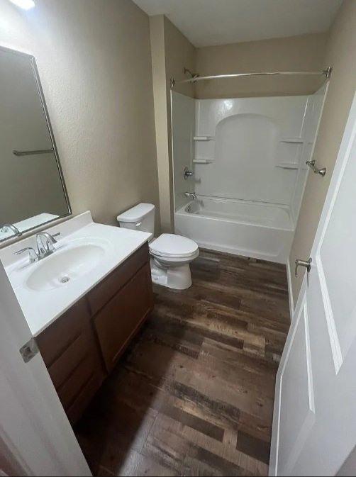 Small bathroom with a white bathtub and shower, a dark wood vanity with a white sink, a mirror, a toilet, beige walls, and wood-look flooring.
