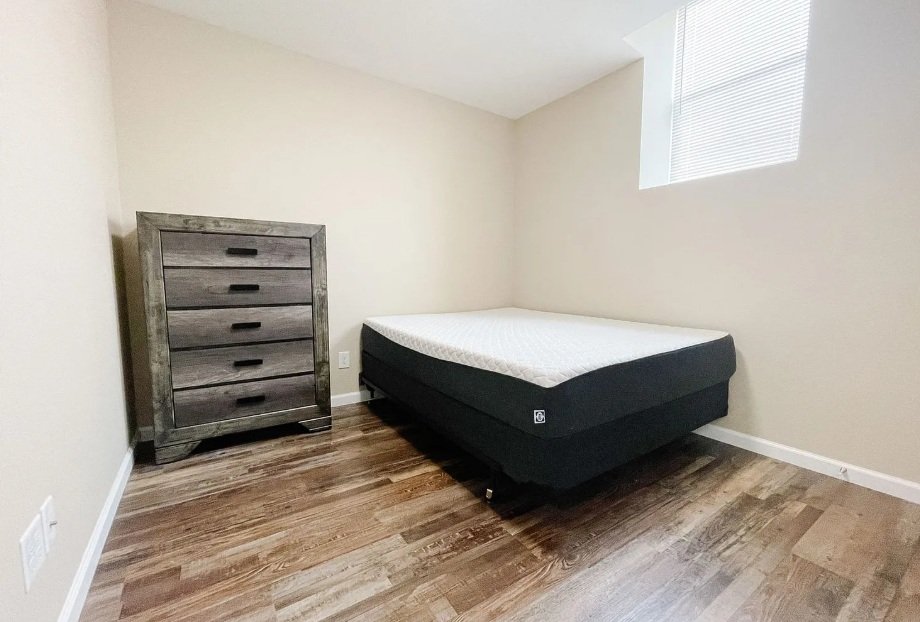 A bedroom with a wooden floor, beige walls, a window with blinds, a bed without bedding, and a gray wooden dresser.