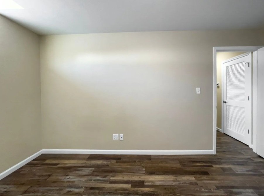 Empty room with beige walls, dark wood flooring, and a doorway with a white louvered door leading to another room.