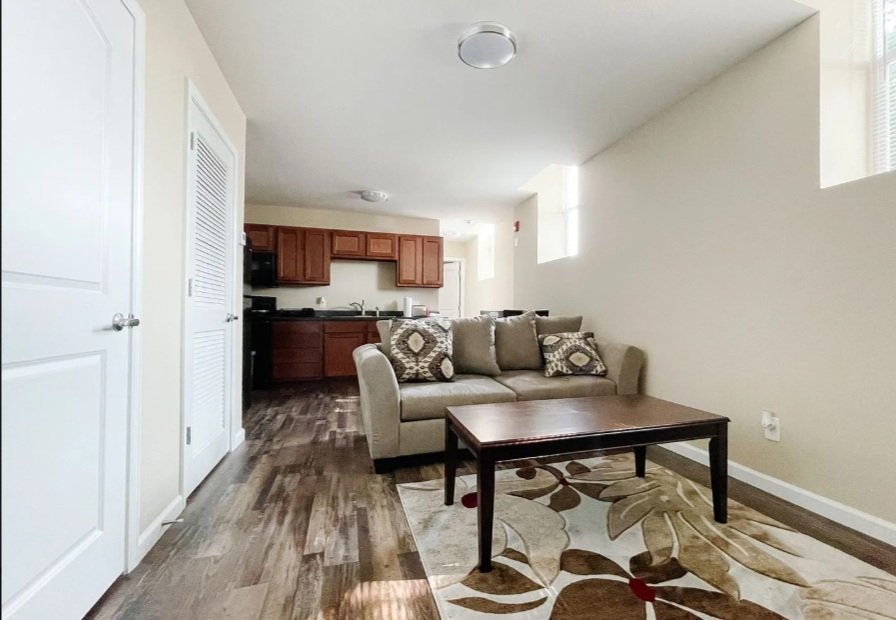 Living room with beige sofa, patterned pillows, wooden coffee table, area rug, and open kitchen with wooden cabinets in the background.
