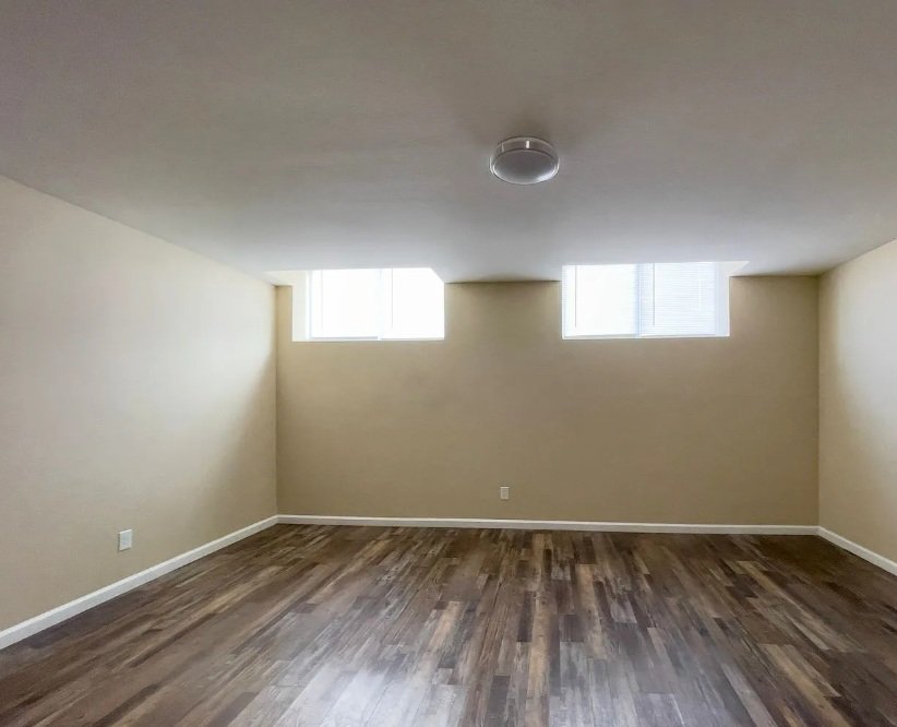 Empty room with beige walls, hardwood floors, and two small rectangular windows near the ceiling, with a ceiling light fixture.