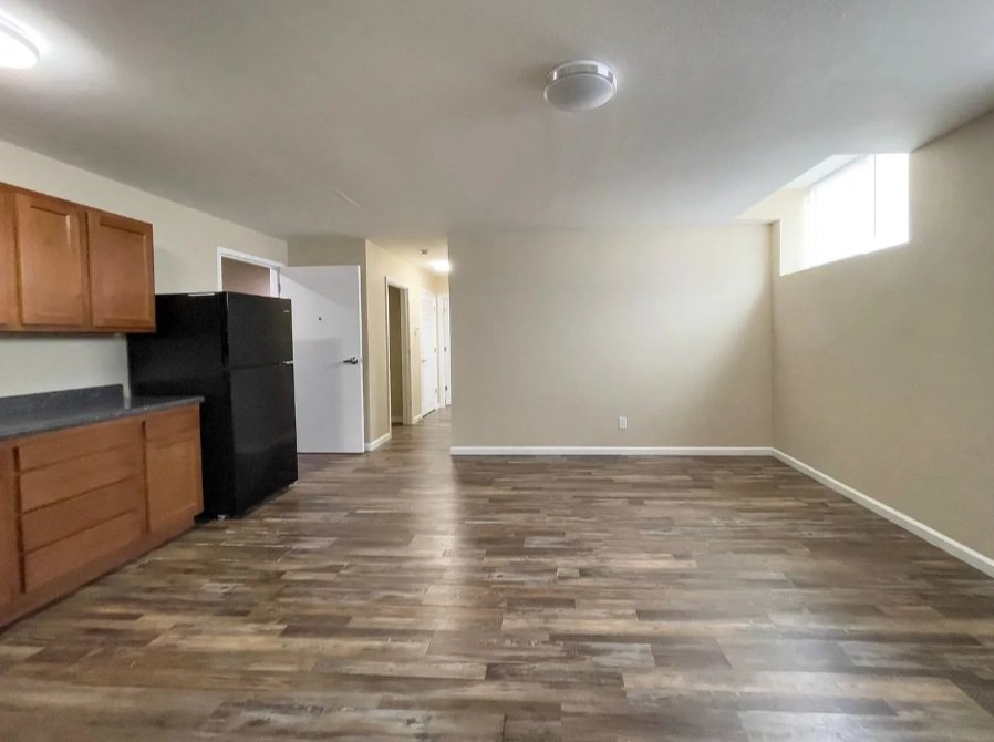 Empty living room with wooden floors, beige walls, a black refrigerator, and a kitchen counter with wooden cabinets, illuminated by natural light from a window.